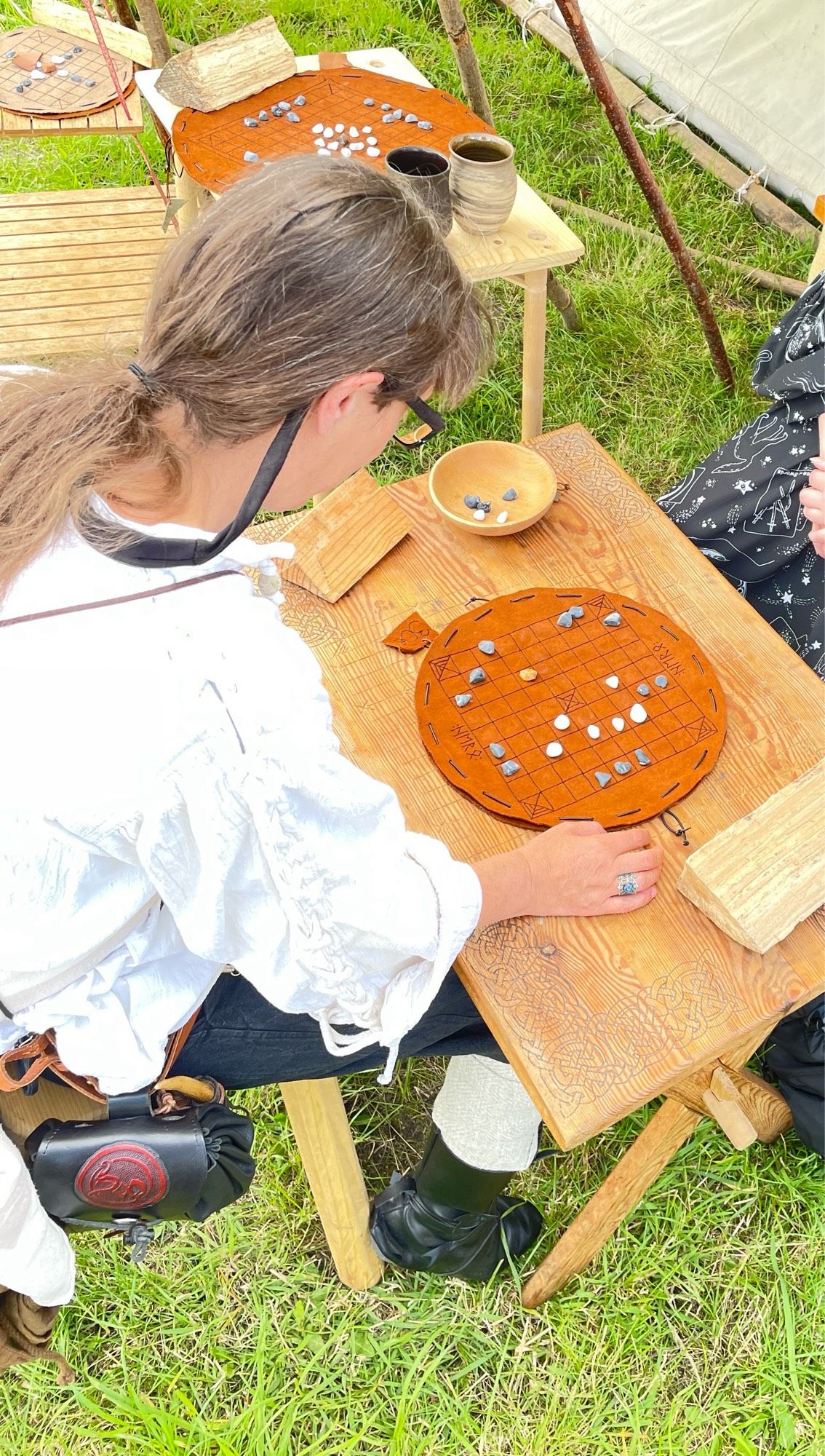 Auf der Wiese am Wikinger Zelt sitze ich am Tisch mit meiner Tochter. Auf dem Tisch 1 rundes Spielbrett aus Leder mit kleinen Steinen in hell und dunkel als Schachfiguren. Wir spielen 1 Runde Wikinger Schach. Bin in Mittelaltertracht : Hose , Hemd , Wadenwickel , Lederboots ,  am Gürtel hab ich meine Tasche u mein Messer.