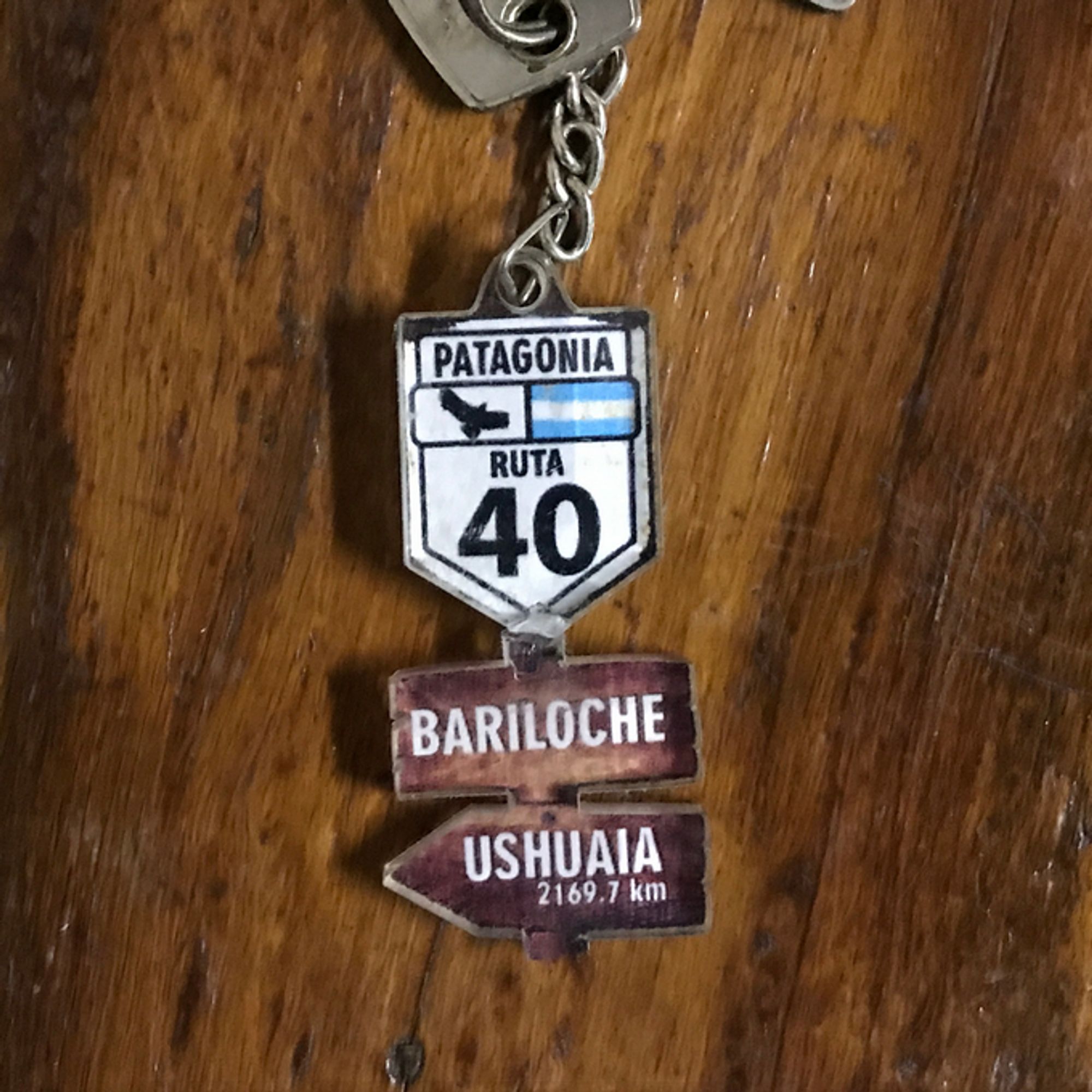 own photo of a resin keychain in the shape of a route sign with the inscription "Patagonia" on top, a condor silhouette on the left, a flag of Argentina to the right, and the inscription "Ruta 40" on the bottom
below, two resin "wooden boards" looking like sign posts say "Bariloche" and "Ushuaia: 2169.7 km"