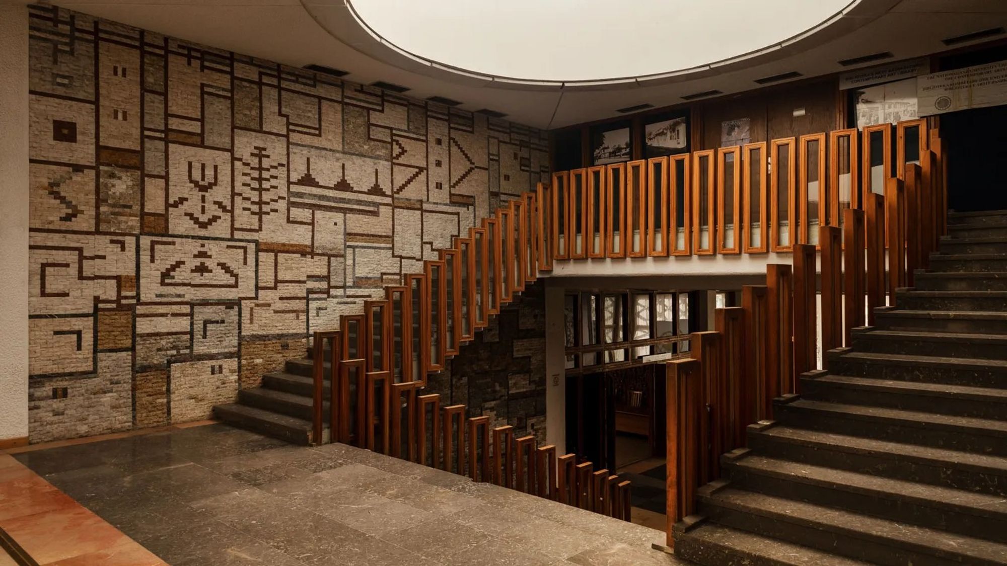 Interior staircase with railing made of repeated tall wooden rectangles and walls decorated with brown and gray mosaic. Photo by Oskar Proctor.