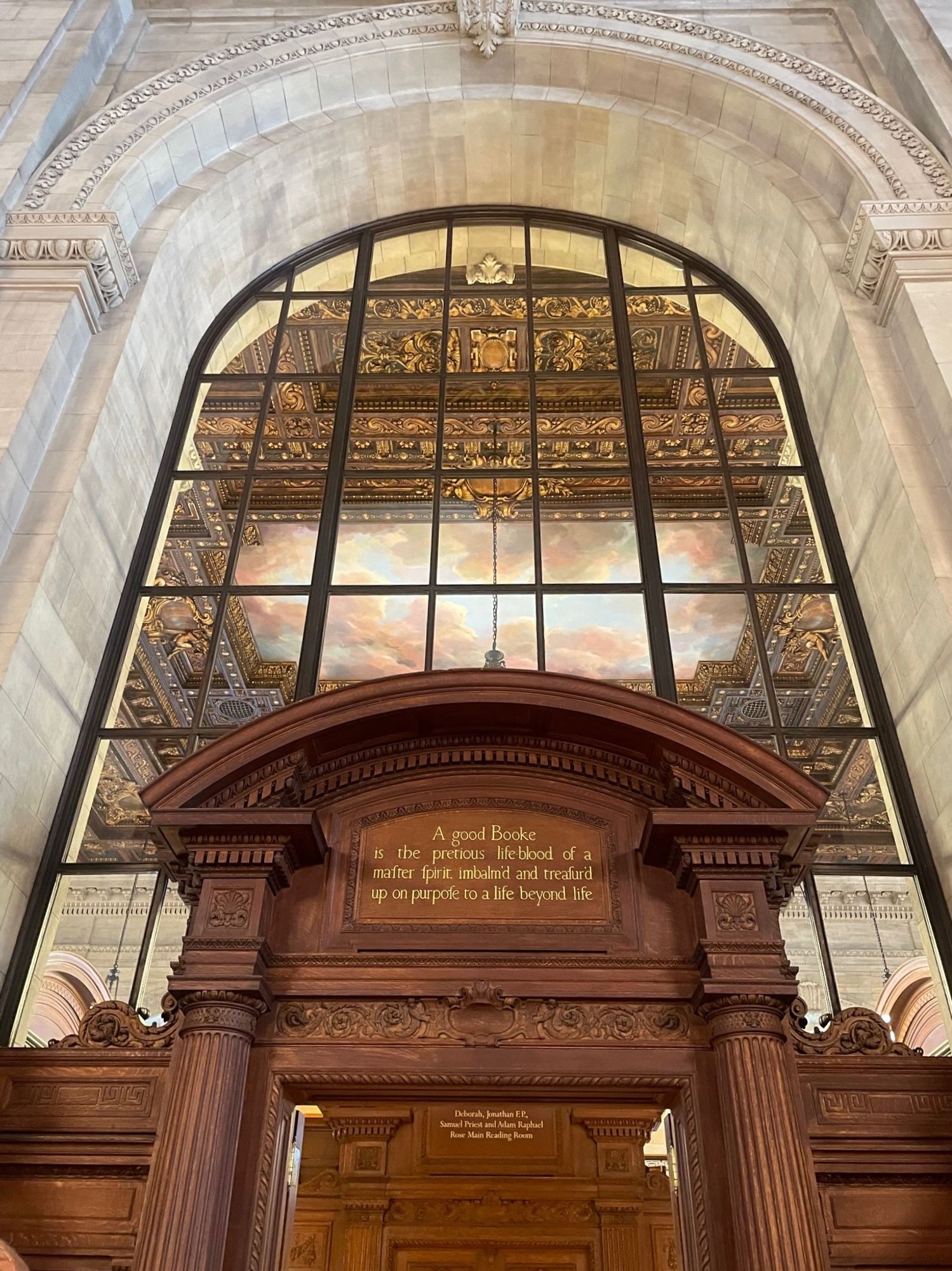 Entrance to the Rose Reading Room at the main New York Public Library. A carved wooden doorway with columns on either side and the following words painted in gold above the threshold: "A good Booke is the pretious life-blood of a master spirit, imbalm'd and treasur'd up on purpose to a life beyond life." Above the doorway is a very tall arched window with many panes letting us glimpse the painted ceiling of the reading room, depicting a blue sky and clouds framed in intricate carved gilt wood.