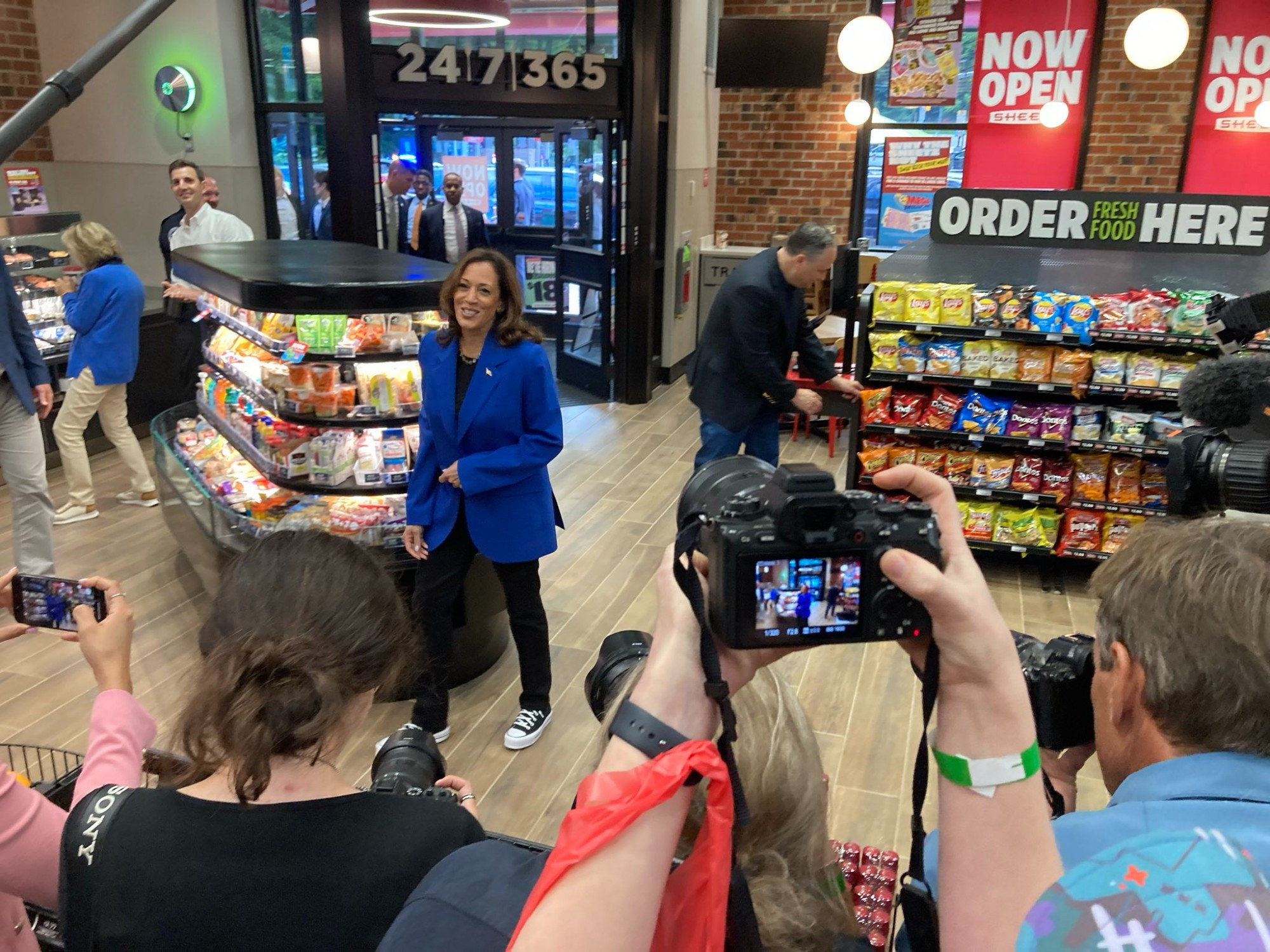 Interior of Sheetz convenience store. Kamala Harris stands in front of a display of refrigerated snacks, making eye contact with a bunch of reporters taking her picture. Photo by Ryan Deto of the Pittsburgh Tribune-Review.