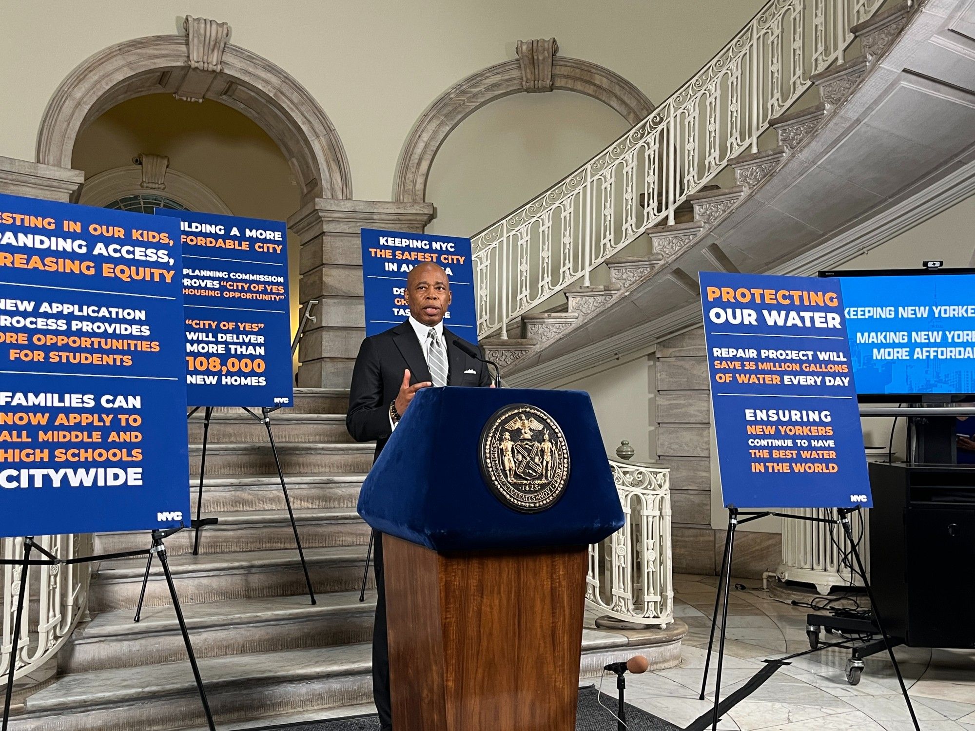 Photo by Craig McCarthy of New York City mayor Eric Adams at a lectern in city hall surrounded by four signs on easels, each sign dense with text in three sections, promoting mayoral accomplishments ("protecting our water," "keeping NYC the safest city in America," "building a more affordable city," etc.)