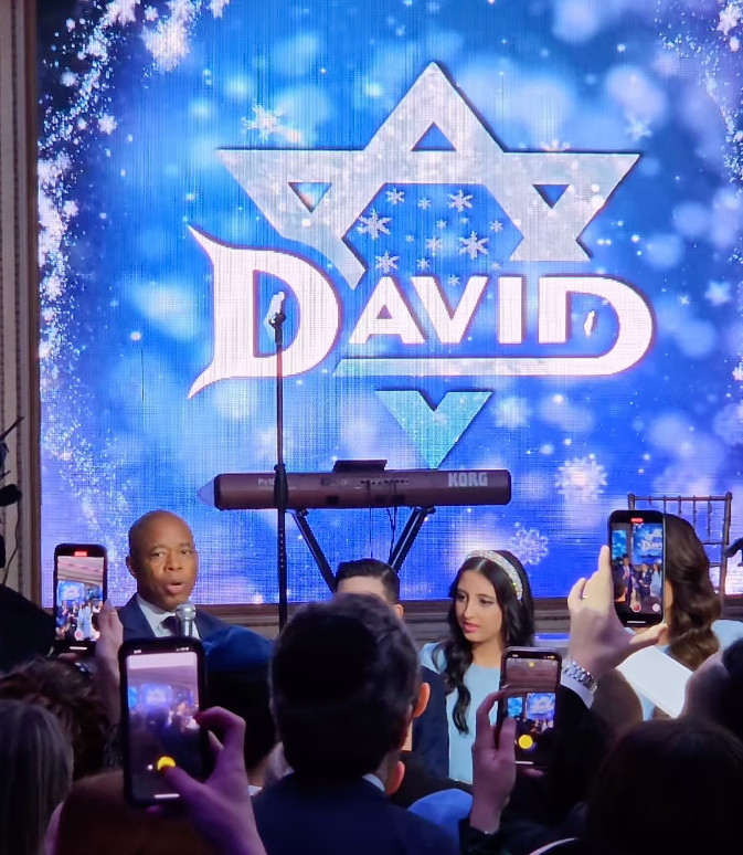 Screencap of Instagram video of Eric Adams speaking at a bar mitzvah. There is an enormous digital background behind him with the name "David" on top of a star of David, with a wintery blue-and-snowflake motif