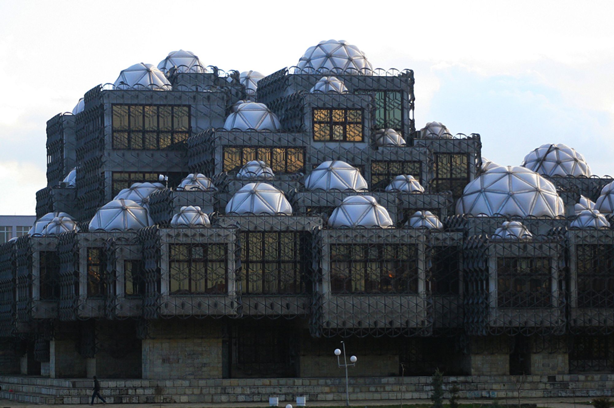 Exterior of library. A brutalist building enclosed by a metal mesh that looks like geometric chain mail, capped by a variety of white domes divided into triangular facets. Photo by Suhejlo CC BY SA 3.0.