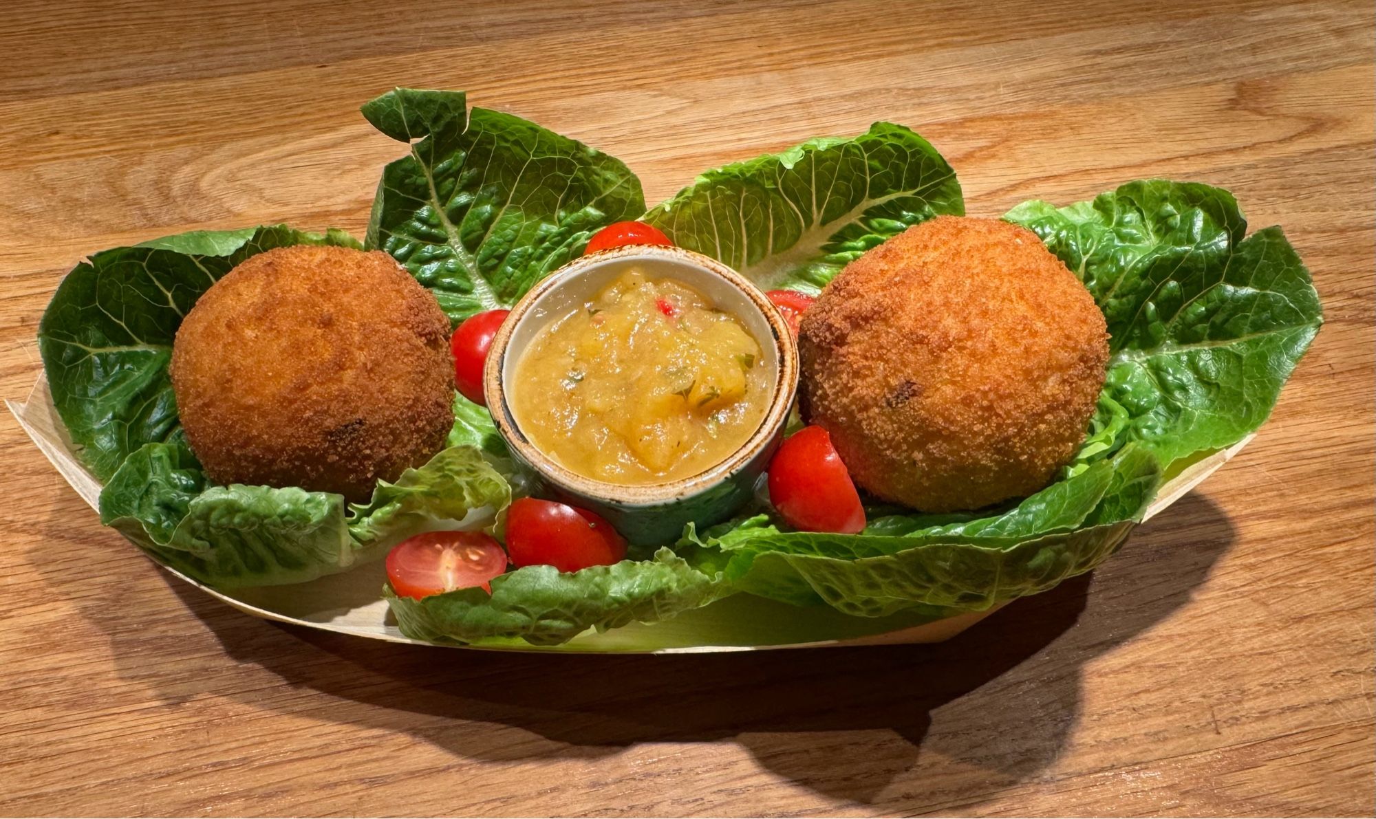 In einer Bananenblatt-Schale liegen auf Salatblättern zwei grosse Bällchen, paniert und goldbraun, in der Mitte dazwischen ein Schälchen mit gelber Tomatensauce, einige halbierte Tomätchen liegen auf den Salatblättern herum.