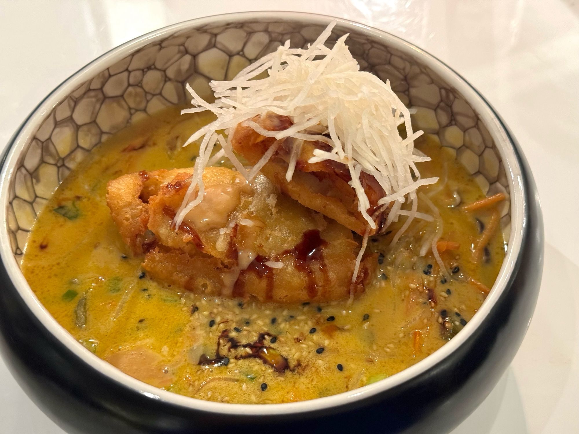 Marmorierte runde Schale mit grünem Gemüse-Curry, obenauf mit Fischfilet im Tempura-Teig. Auf dem Fisch marinierte Rettich-Streifen.