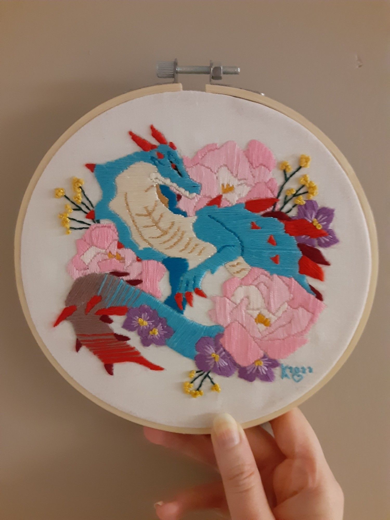 A Lagiacrus (a blue electric sea dragon from monster hunter) surrounded by pink peonies and some small purple and yellow flowers all done in embroidery