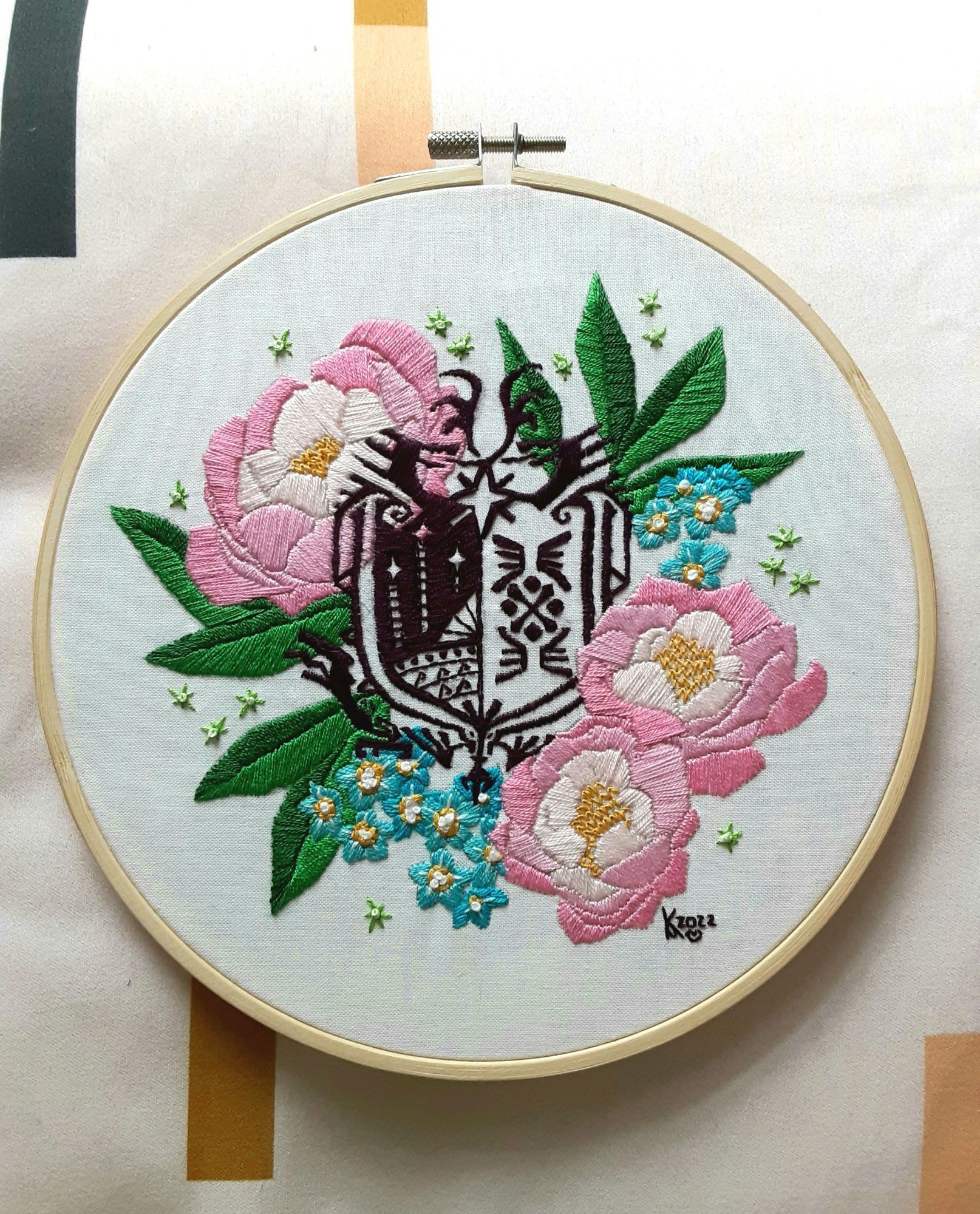 An embroidery piece of the monster hunter world commission symbol surrounded by pink peonies and forget me nots