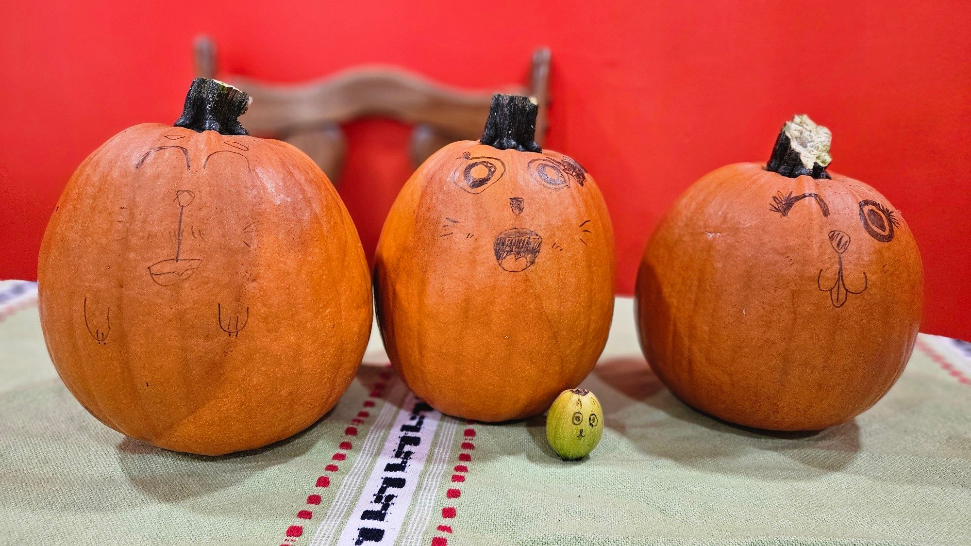 3 big pumpkins and one tiny green pumpkin on a table (green tablecloth, orange wall in background). The pumpkins have faces drawn on them.