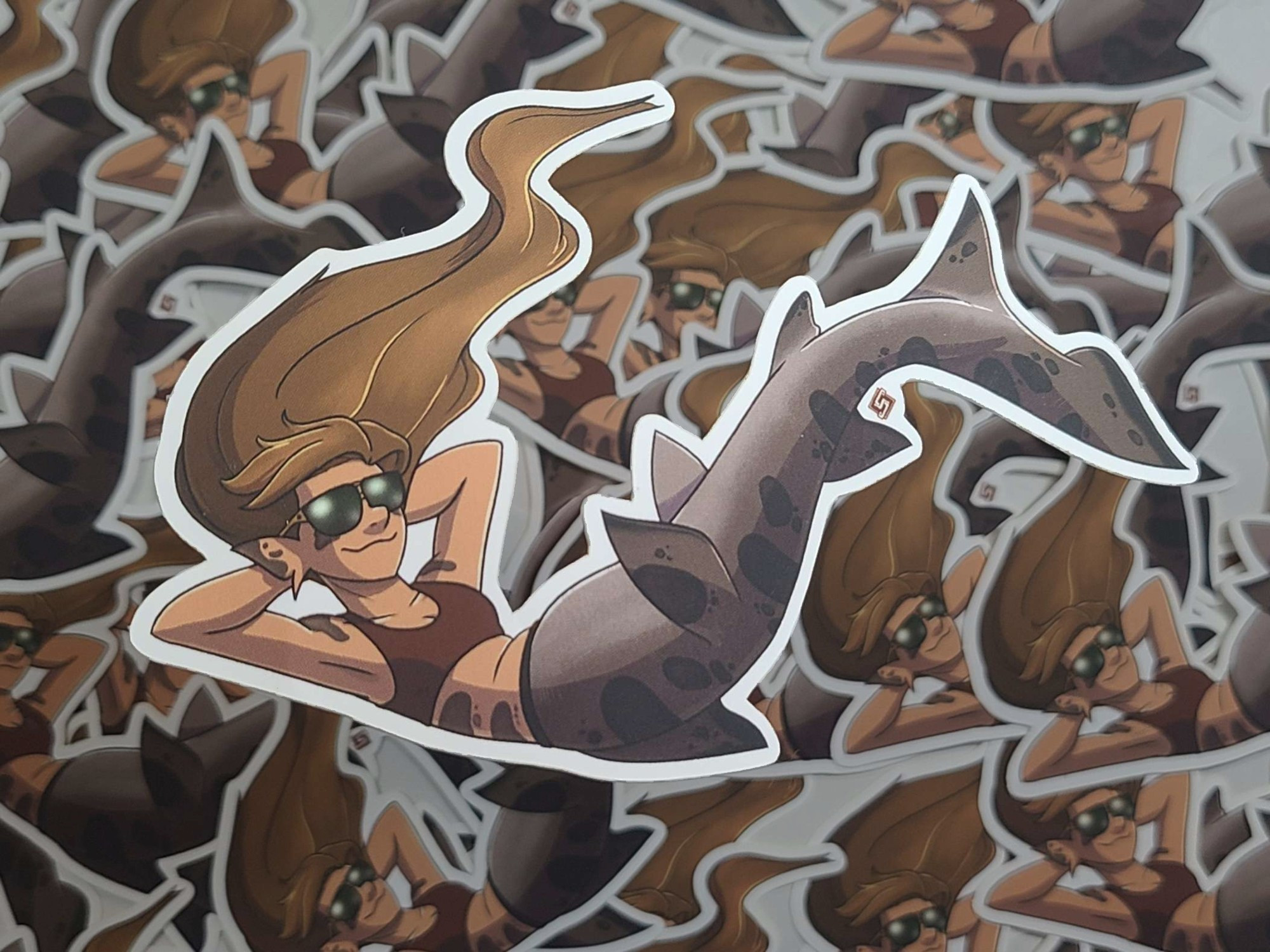 A sticker of a leopard shark mermaid. She has long brown hair and a pair of sunglasses on. She's in a reclining pose.