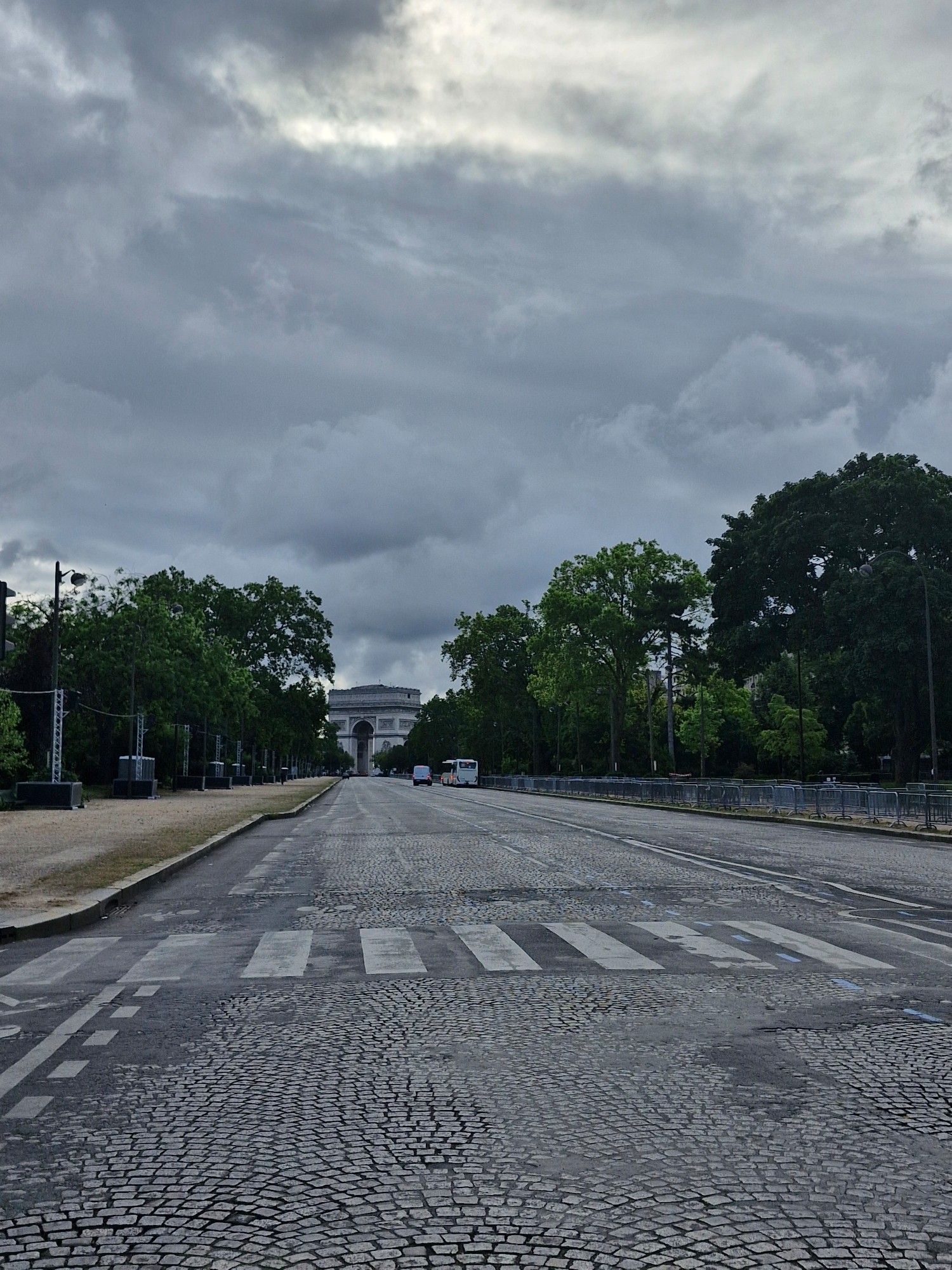 Avenue Foch vide de voitures et de feux de circulation, vue sur l'arc de triomphe. L'avenue pavée est bordée de trottoirs en sable et d'arbres en feuilles et de barrières sur le trottoir droit. Ciel gris et nuageux.