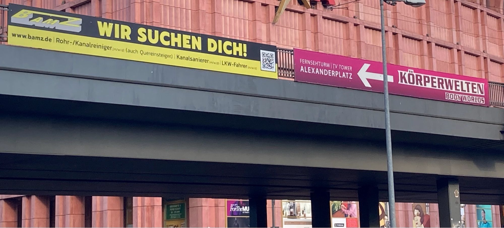 Eine Brücke mit zwei großen Werbebannern. Auf einem steht „Körperwelten“ und ein Pfeil, der auf das andere Banner zeigt. Auf dem zweiten steht „Wir suchen Dich!“