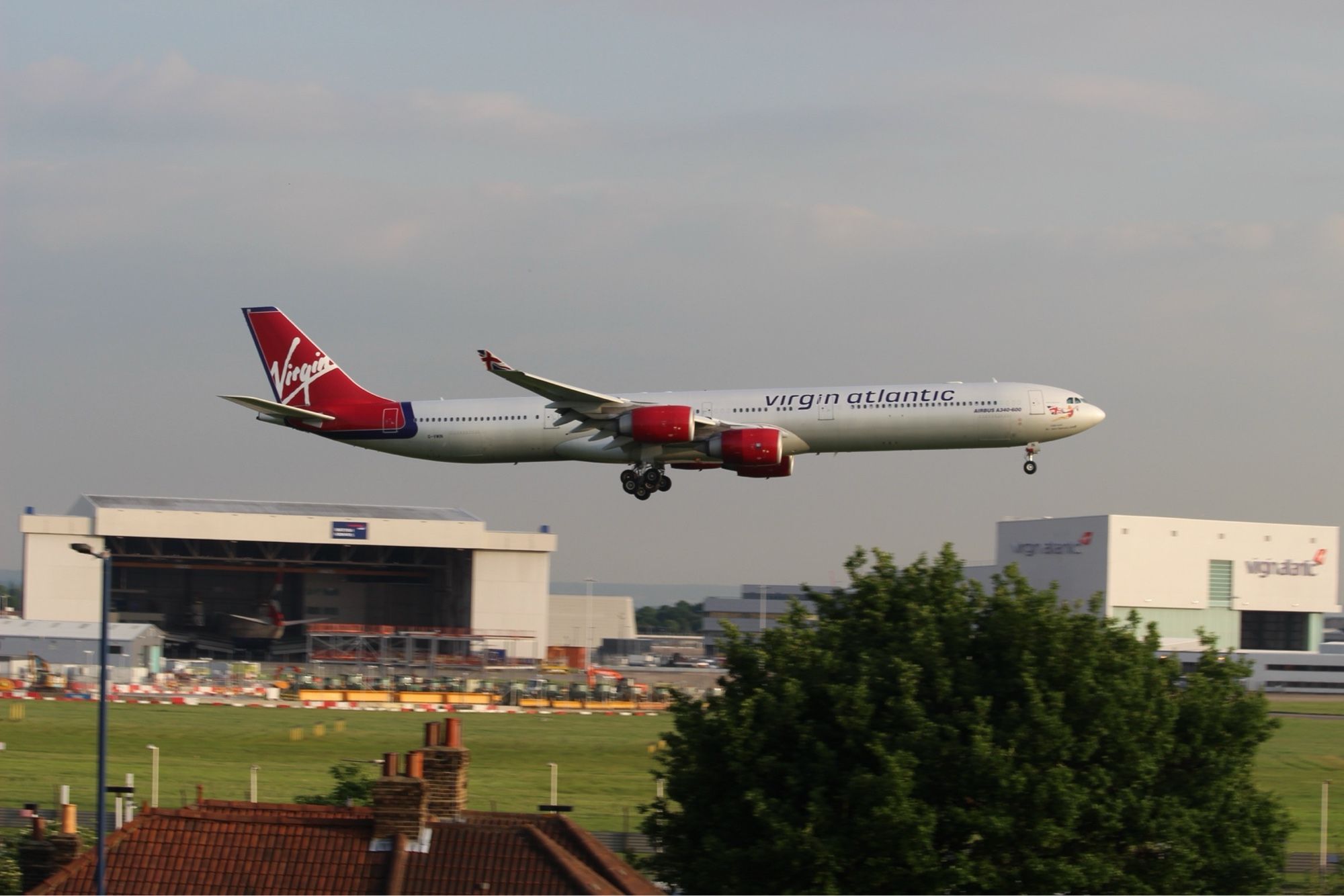 A Virgin Atlantic A340 in purple band livery lands over the BA and VS hangars at Heathrow