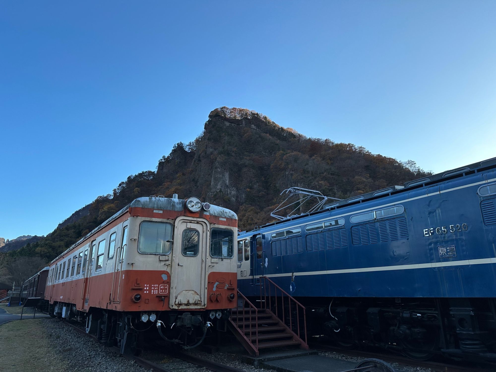 Three vintage Japanese trains sit outside on some tracks in a park, with a mountain in the background.