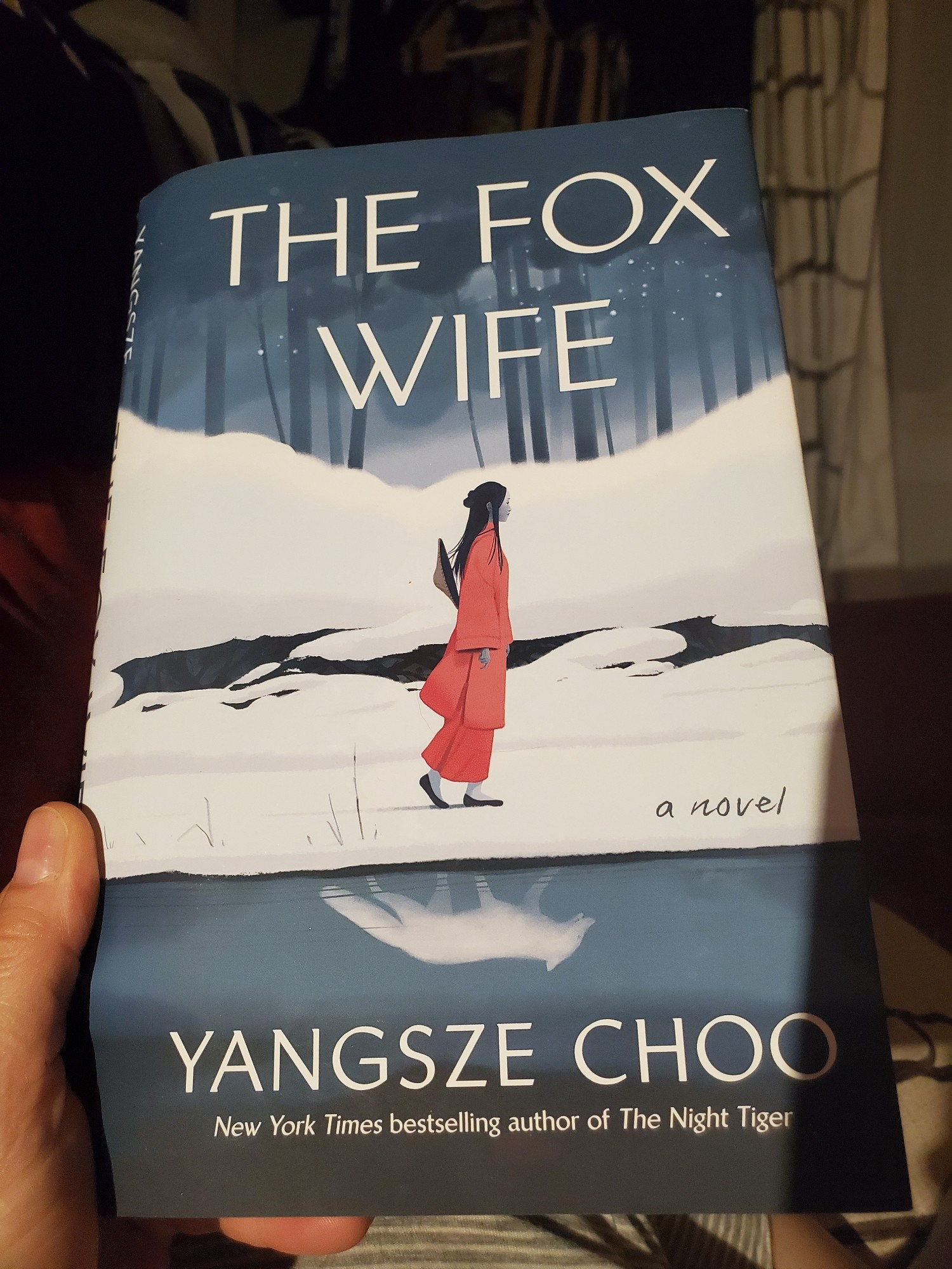 a hand holds a hardcover copy of The Fox Wife by Yangsze Choo. There is all illustration of a woman dressed in red walking in a snowy blue and white landscape.