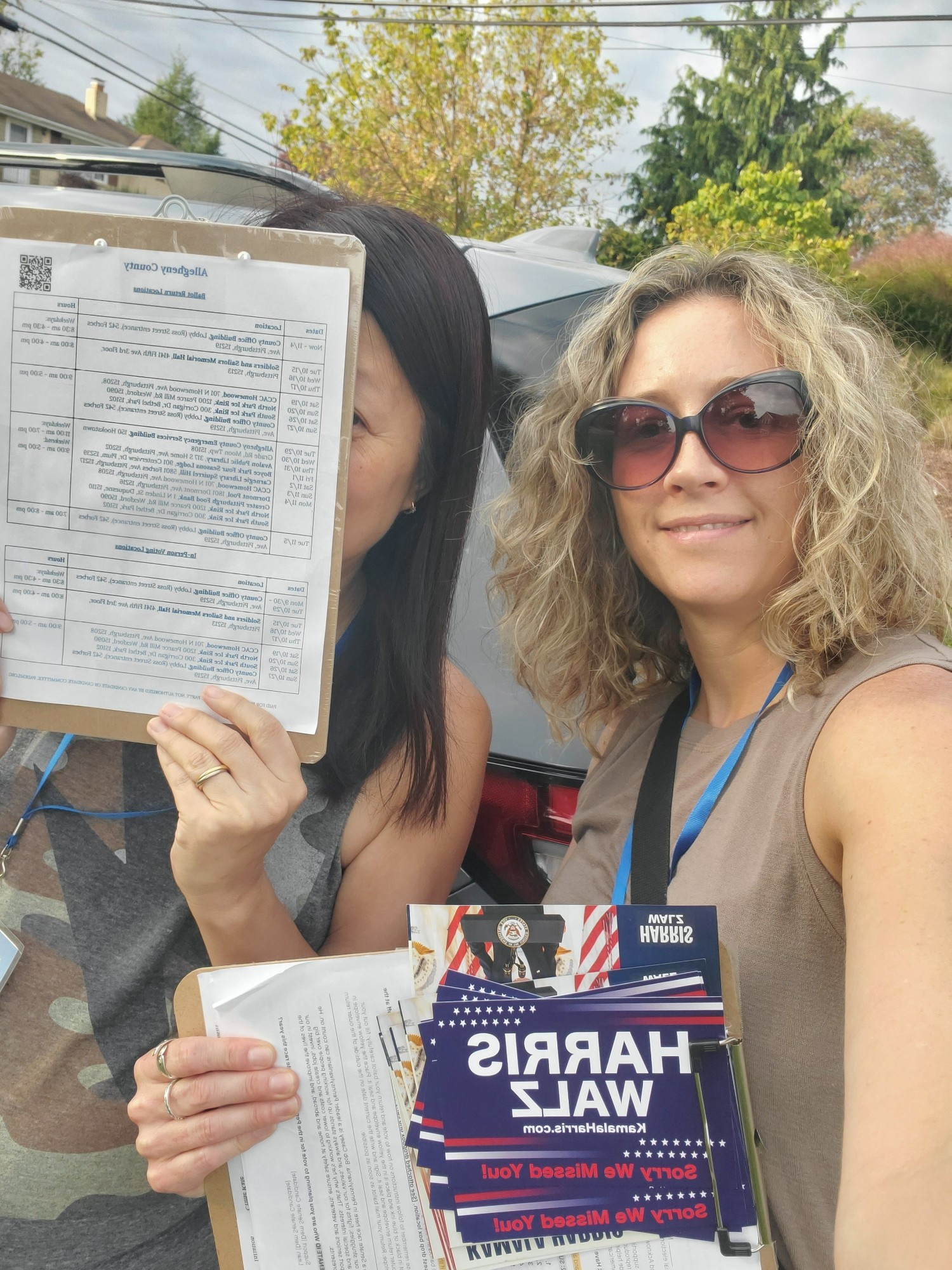 selfie of two women holding clipboards and Harris/Walz lit. the woman on the right is blonde and wearing sunglasses and a sleeveless shirt, the woman on the left is obscuring her face behind a clipboard