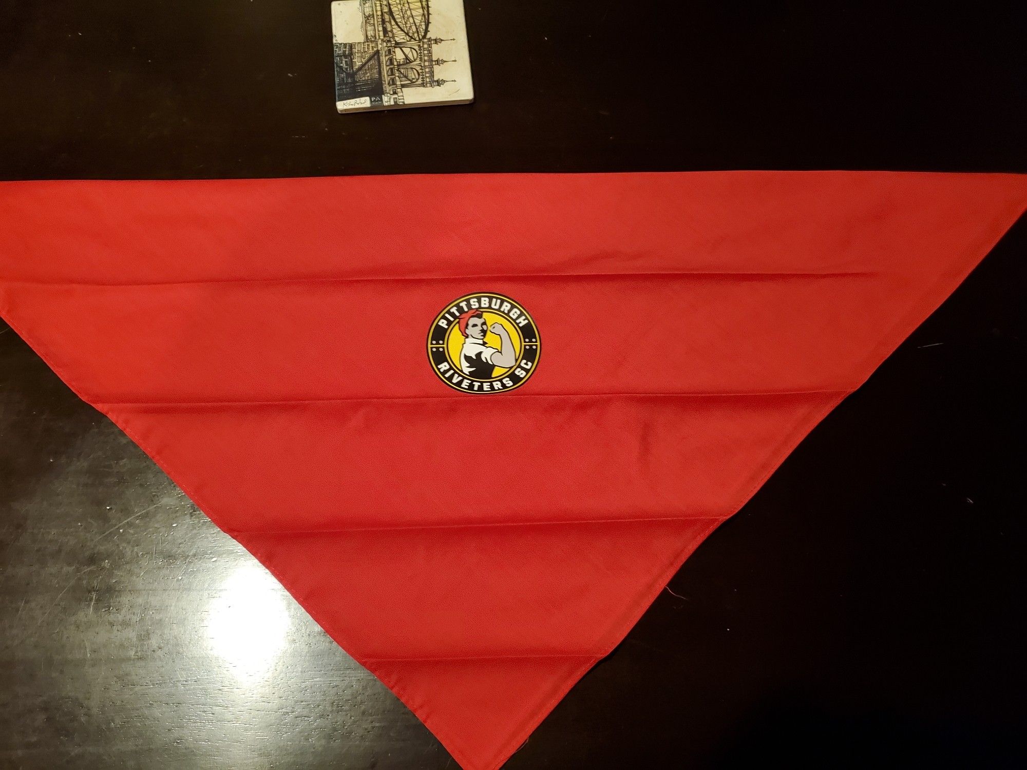 bright red bandana folded in half to be a triangle with the pittsburgh riveters soccer club logo in the middle, it features Rosie the riveter