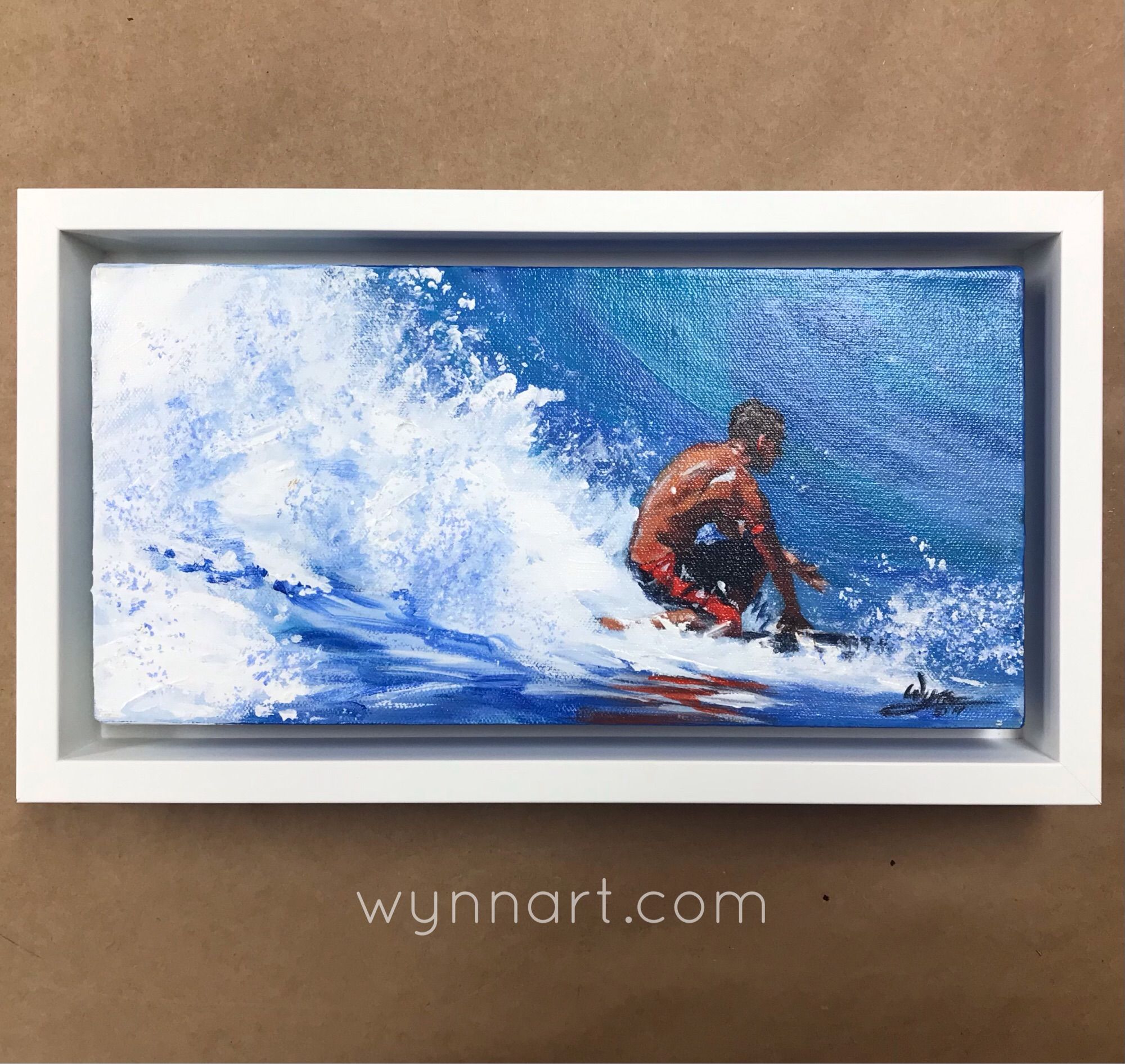 Photo of acrylic painting of surfer surfing on one knee with white wave behind him. Painting has a white float frame.