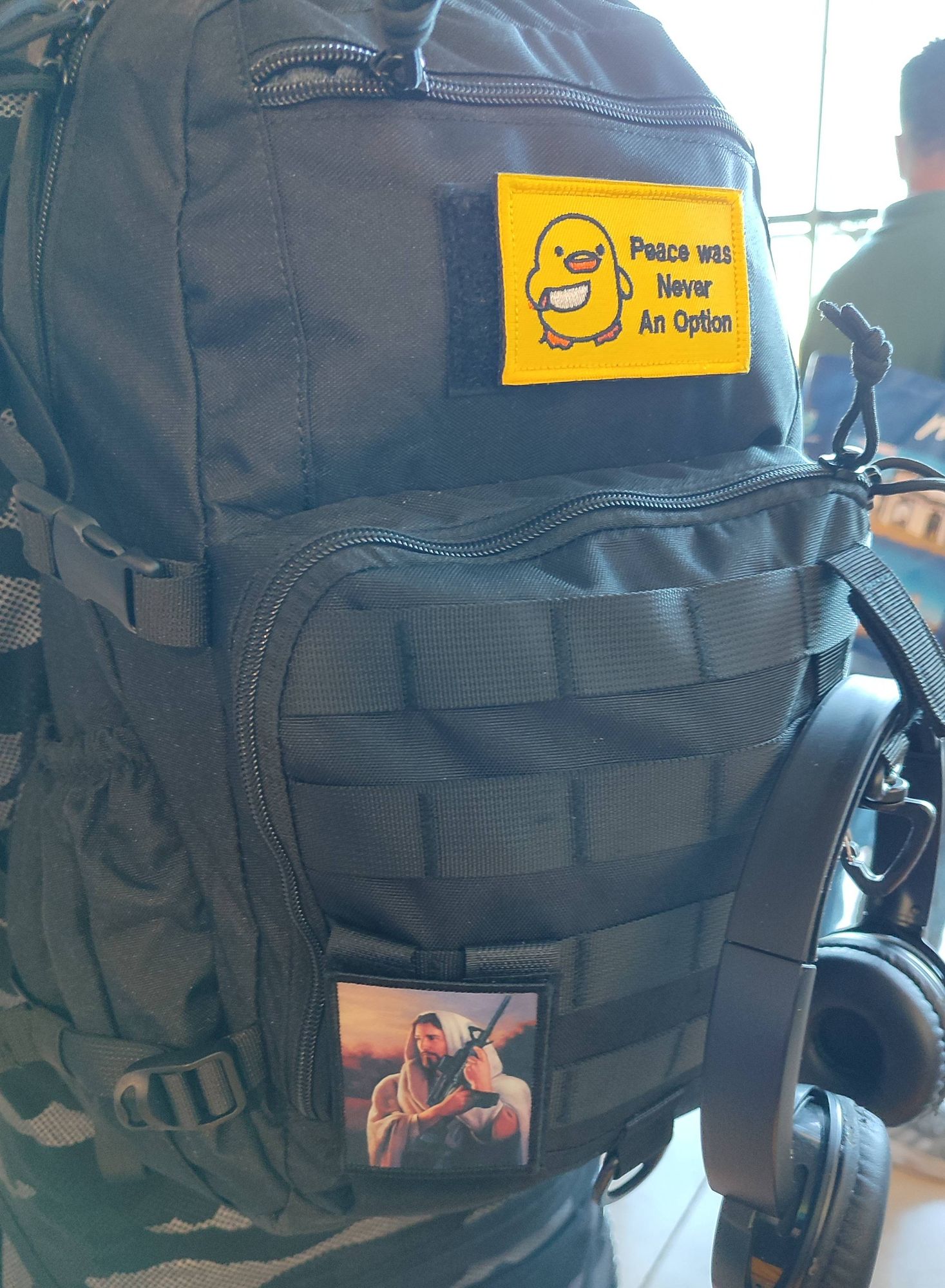 Mochila táctica con dos parches. En la parte superior centrado un parche amarillo con un simpático patito con un cuchillo en el que se puede leer "peace was never an option", "La paz nunca fue una opción".
En la parte baja a la izquierda bajo un bucolico atardecer un Jesucristo posa agarrando un M16.