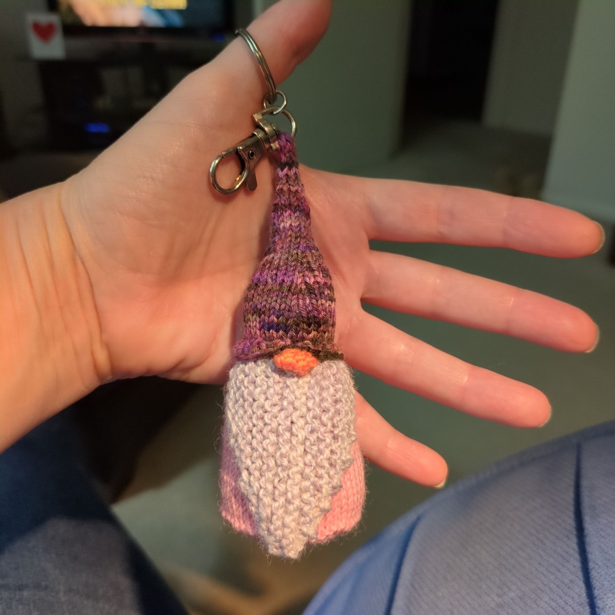 A small handknitted gnome in shade of of pink/purple.
The hat is attached to a keyring.
The keyring hangs from my thumb so my hand is in the background.