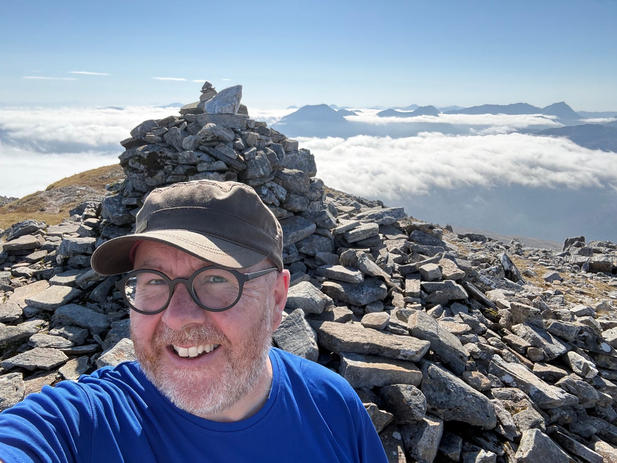 Me on the summit, cap and blue top, in the sun, clouds and peaks behind 