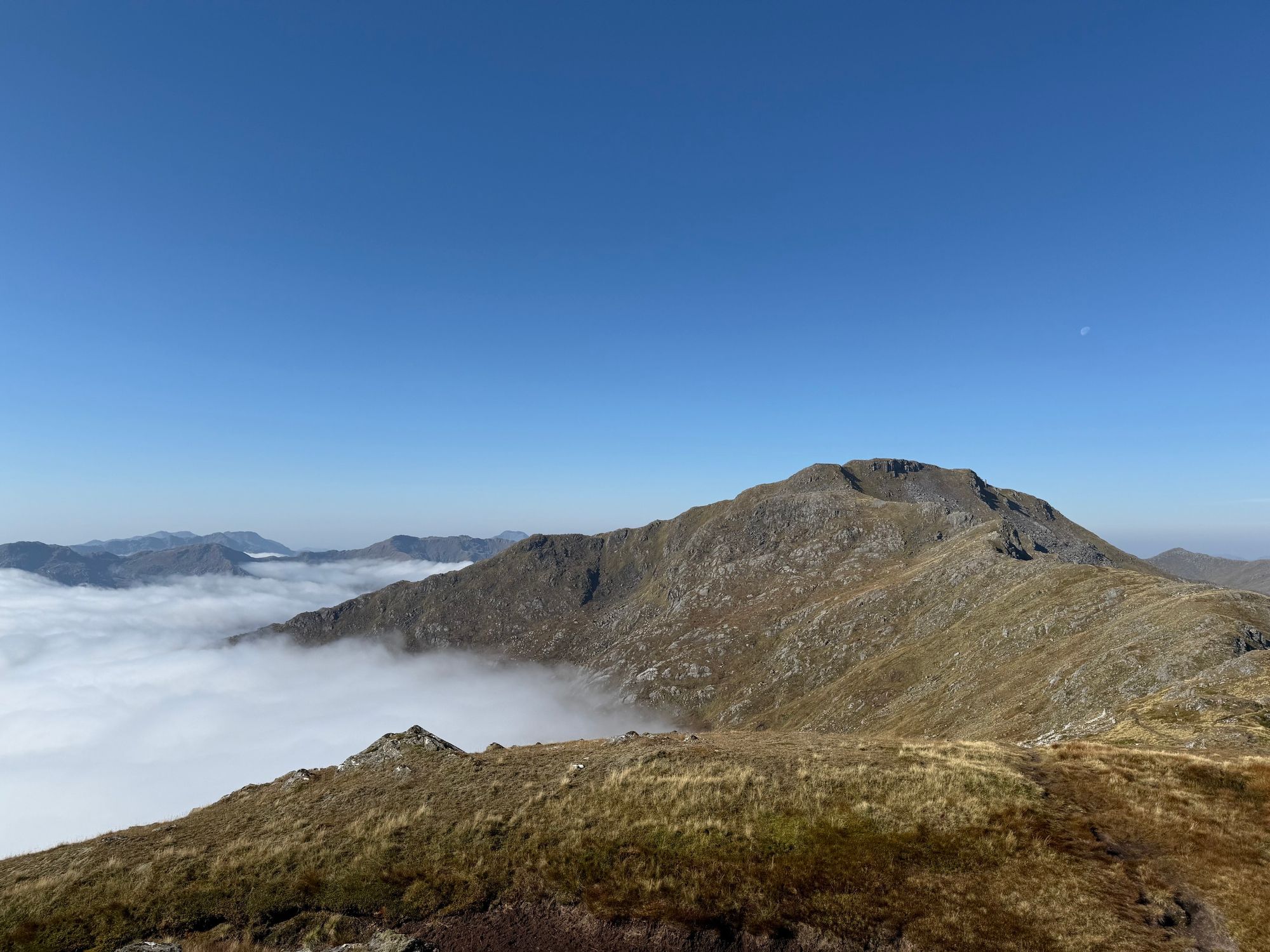 Summit of Sgurr a’Mhaoriach in the distance with the moon out, blue sky, but a carpet of cloud below