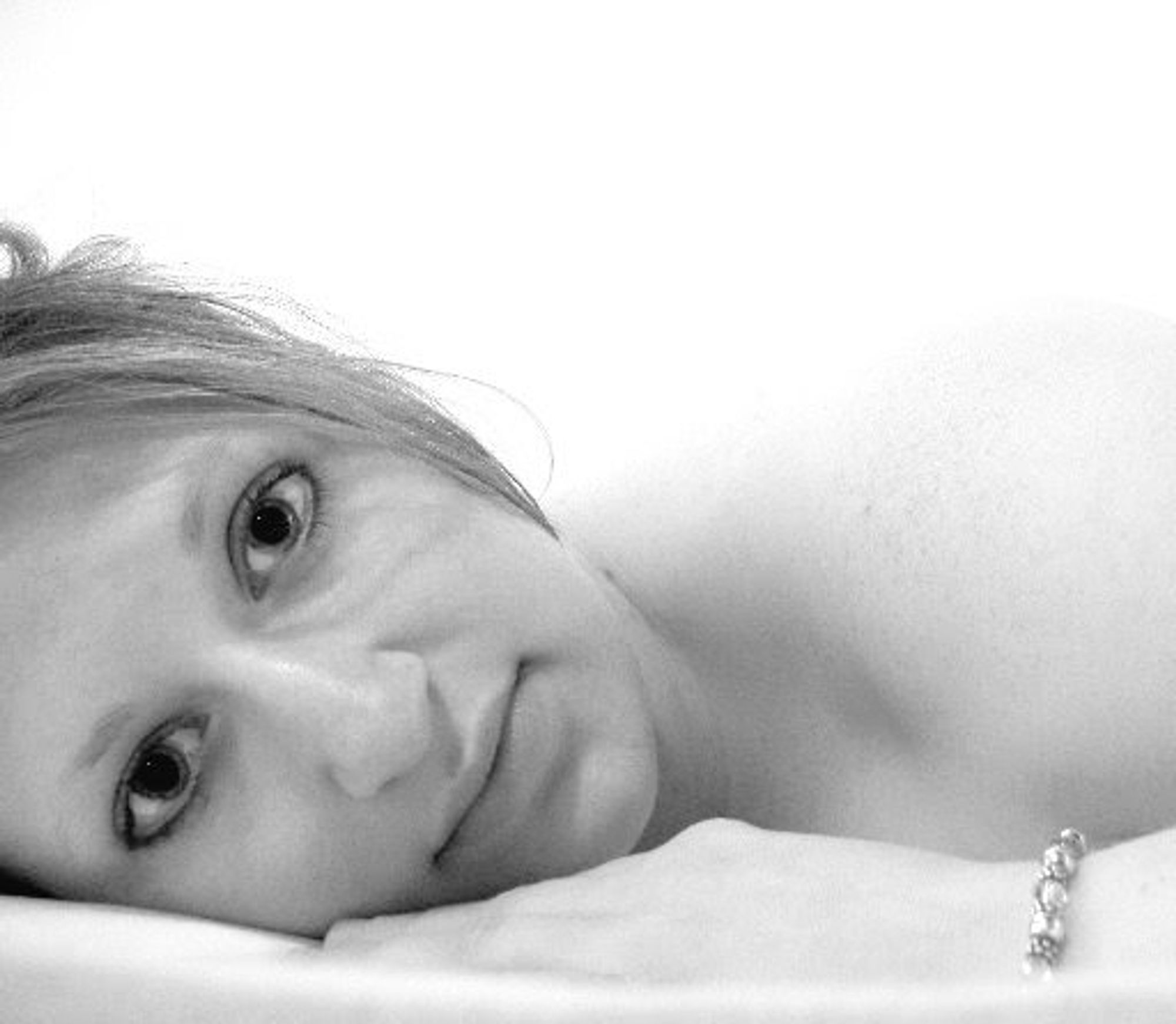 Black and white photo of a white woman with her hair framing her face, laying naked in a bed. She is laying on her stomach and has one hand up by her cheek, while a slight smile plays on her lips.