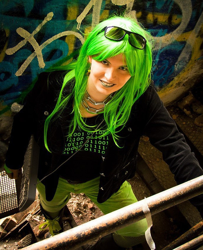 A woman with long, bright green hair, wearing sunglasses on top of her head, stands on a metal stairway in front of a graffiti-painted wall. Rubble lies around on the stairs, a metal bar crosses the lower foreground in front of her. 
She's wearing bright green trousers and lipstick, a black top with bright green binary code printed on, and an old keyboard. A heavy metal chain, wrapped around her neck twice, works as he jewellery.