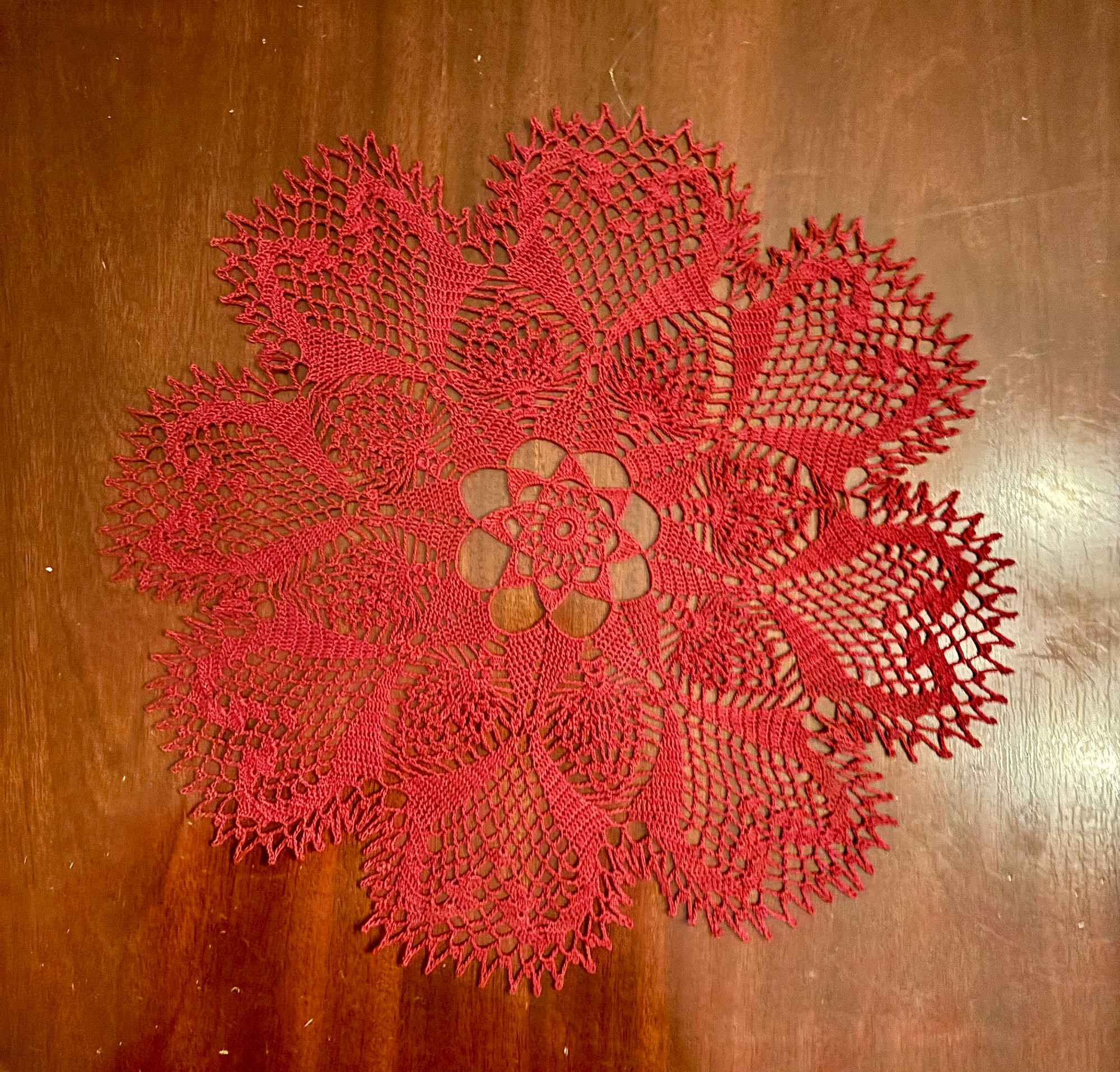 a crocheted red lace doily, around a foot in diameter, on a light brown wooden table