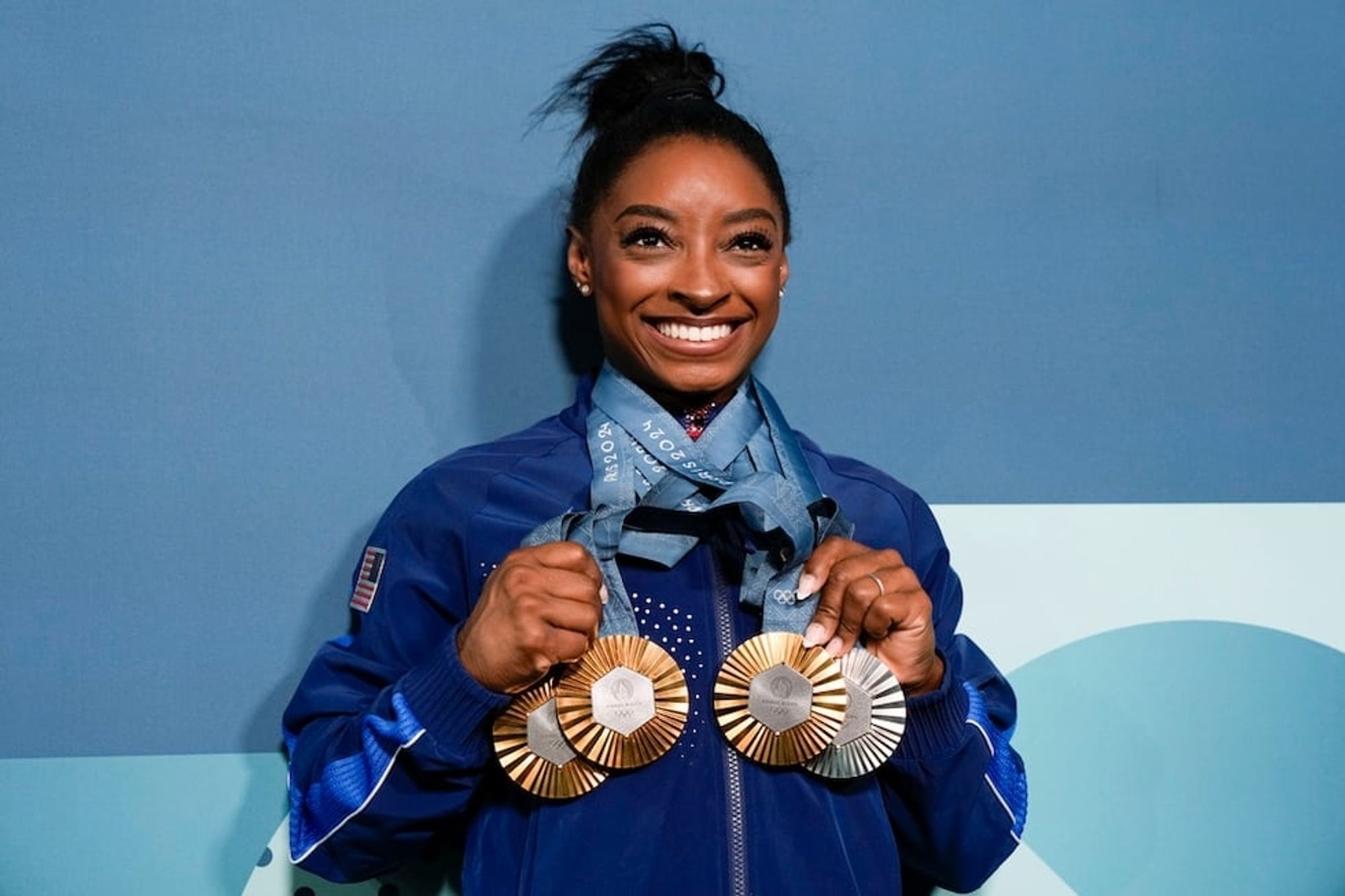 Simone Biles poses for a photograph. Smiling ear to ear she holds up the three gold and one silver Olympic medals she earned at Paris, which are all draped around her neck.