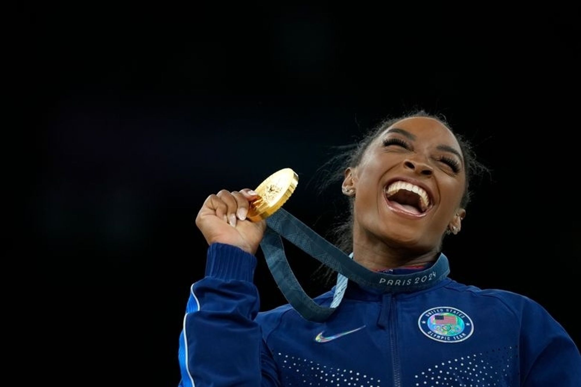 Simone Biles at the Paris Olympics. With an absolutely enormous grin she raises her gold medal up in celebration.