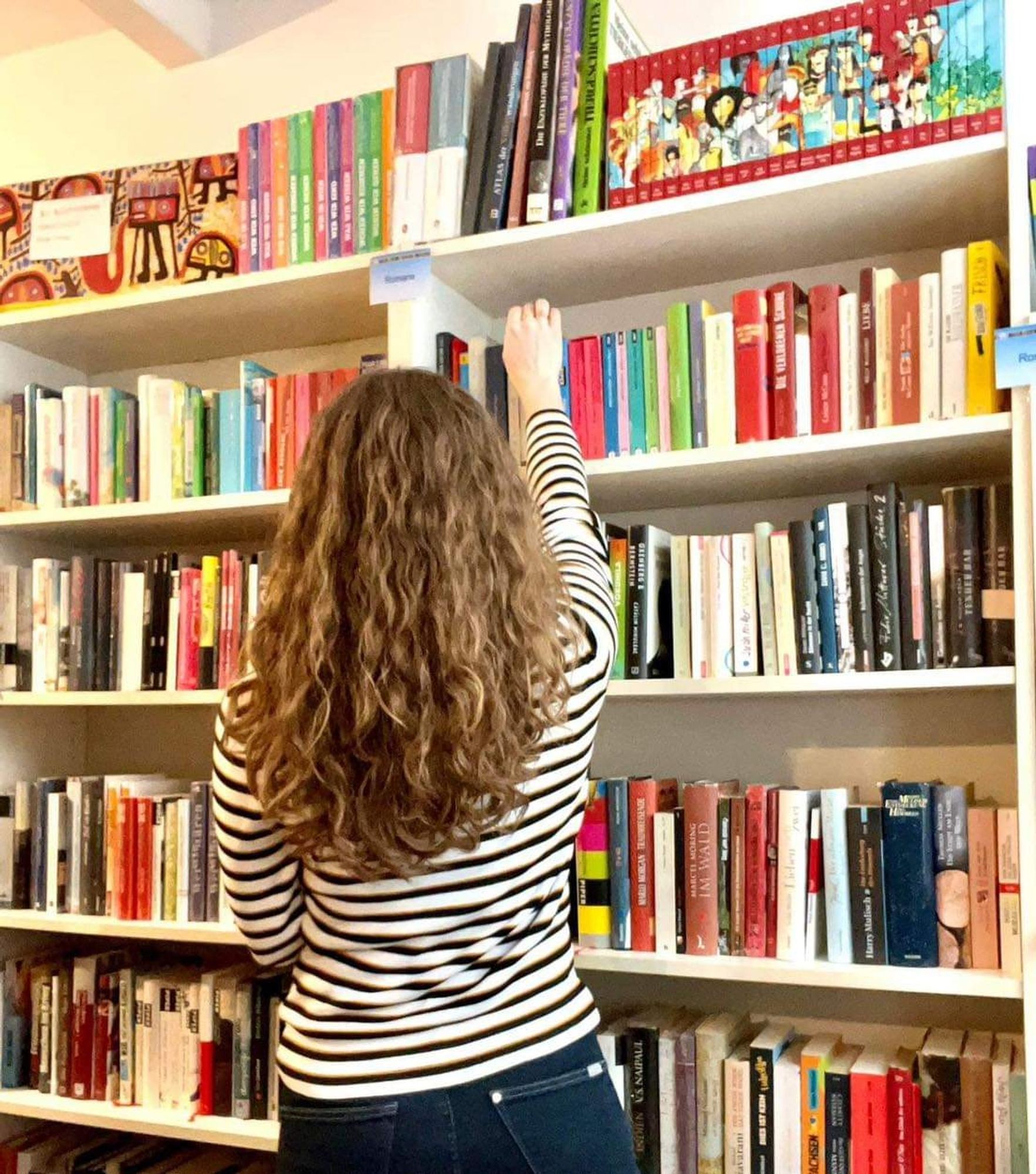 Eine Frau mit langen, lockigen Haaren steht vor einer Wand voller Bücherregale und holt ein Buch aus dem oberen Fach