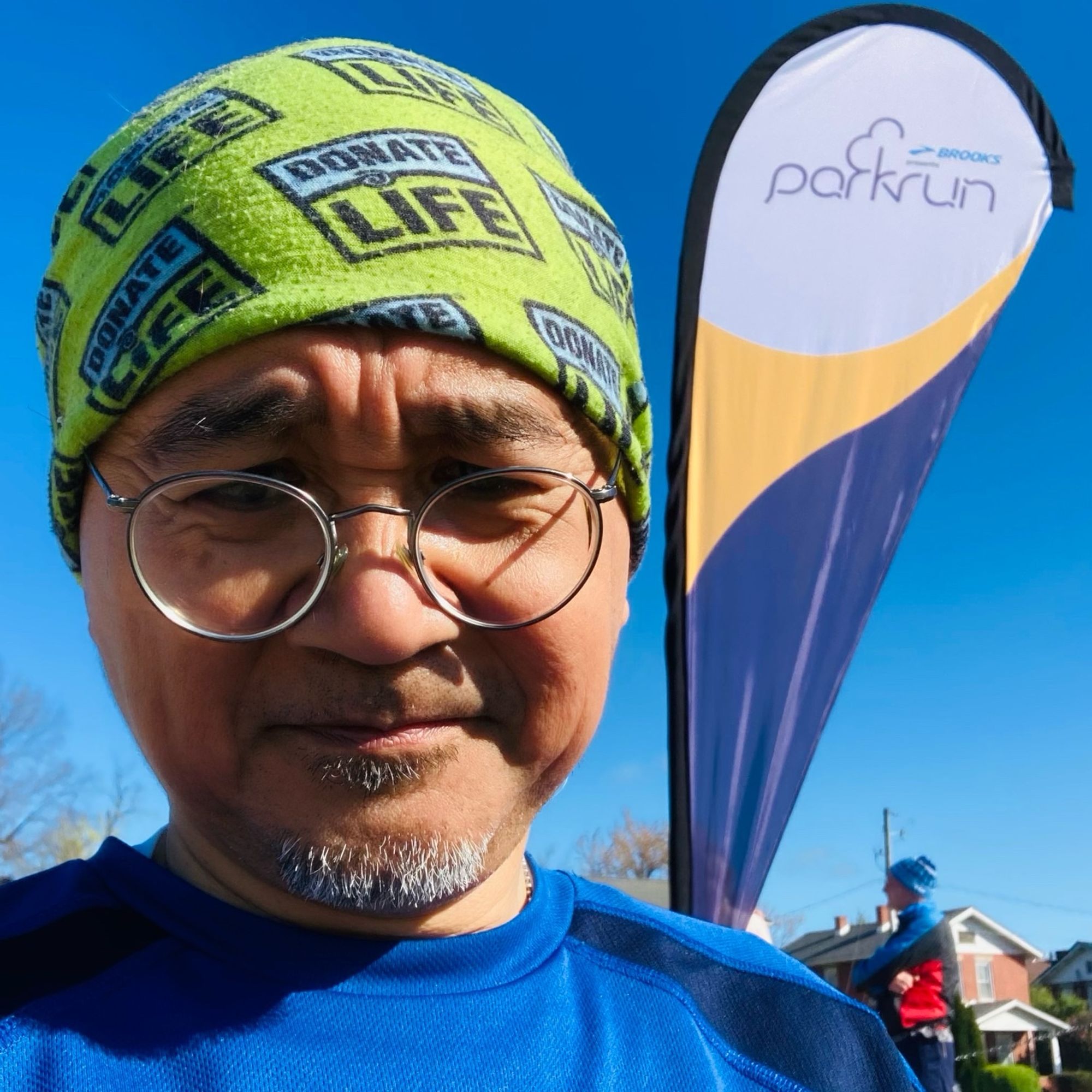 Man wearing “Donate Life” gaiter on his head to promote organ donation. In background is parkrun 5k banner.