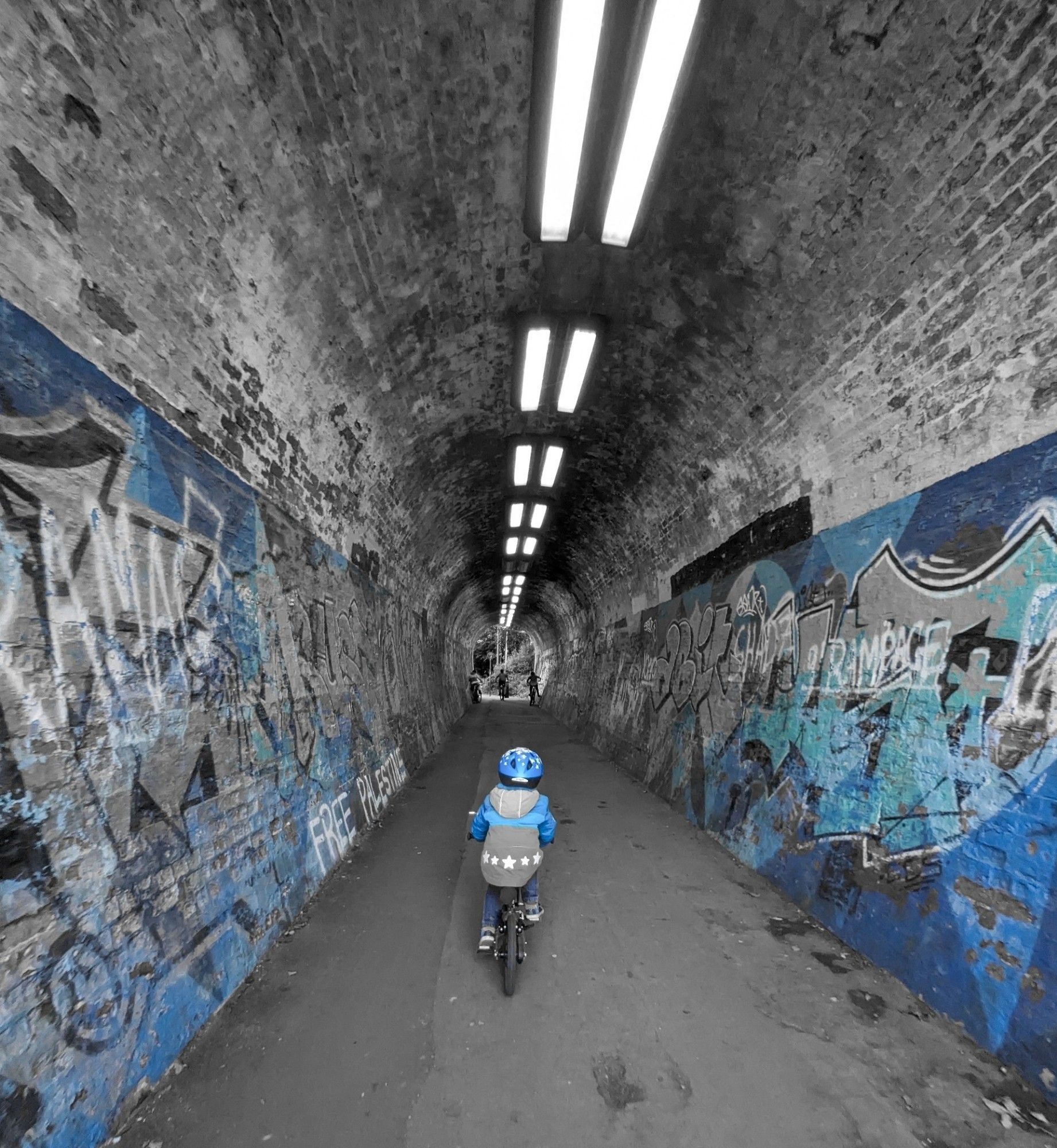 Black and white photo with blue colour pop of small boy riding a pedal bike through a graffitied tunnel