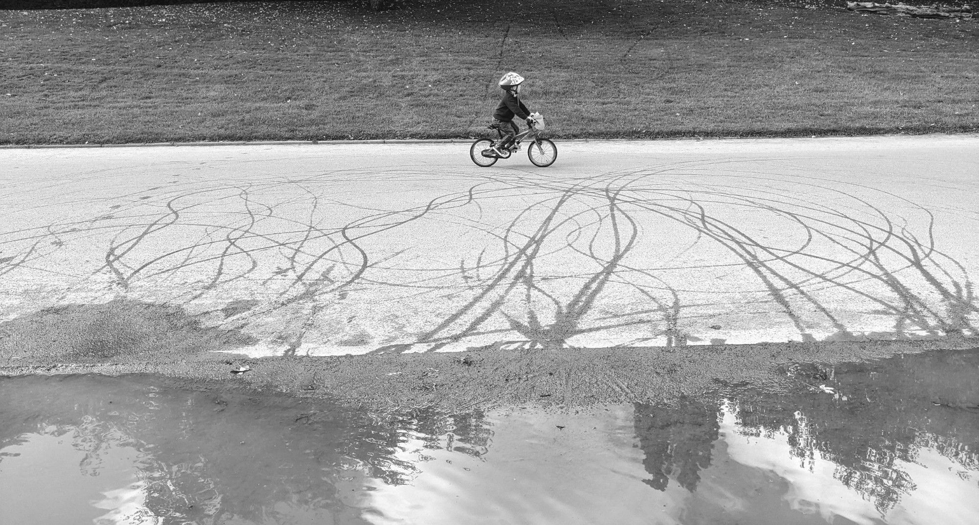 Black and white photo with a large puddle in the foreground with trails left by wet tyres coming out of it nd at the 'artist' (a little boy on a tiny pedal bike) is in the background riding past leaving his mark.