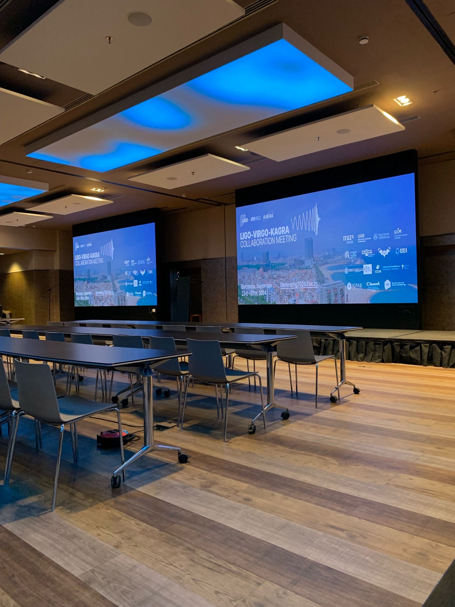 Image of an empty conference room with screens for presentation lit up with the conference logo.