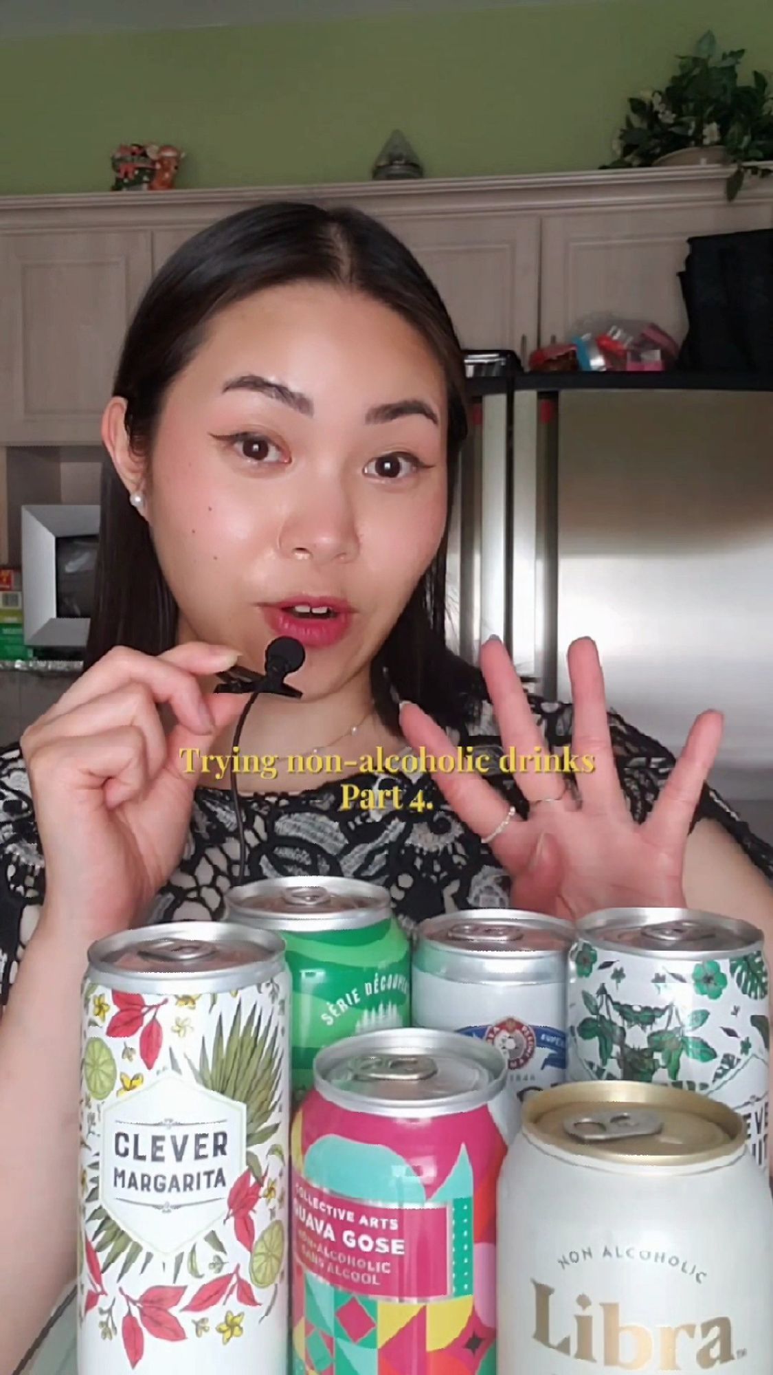 A woman in a kitchen with her right hand holding a microphone to her mouth and her left hand is showing 4 fingers. There is yellow text in her upper chest area that says, "Trying non-alcoholic drinks Part 4". In front of her are 6 cans of non-alcoholic drinks.