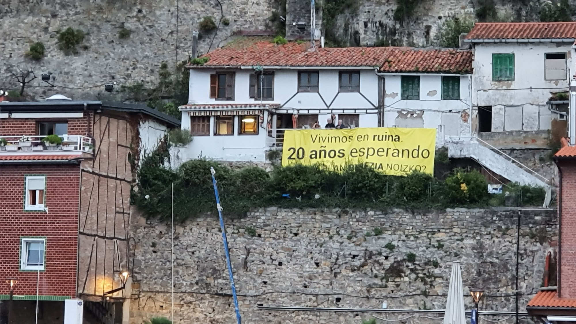 Vor einem Haus hängt ein gelbes Spruchband mit der Aufschrift: 
Vivemos in ruina 20 años esperando
Etwa: wir leben 20 Jahre in einer Ruine und hoffen.
Das Haus ist an eine Felswand gebaut, links davon ein Fachwerkhaus, im Vordergrund eine Steinmauer 