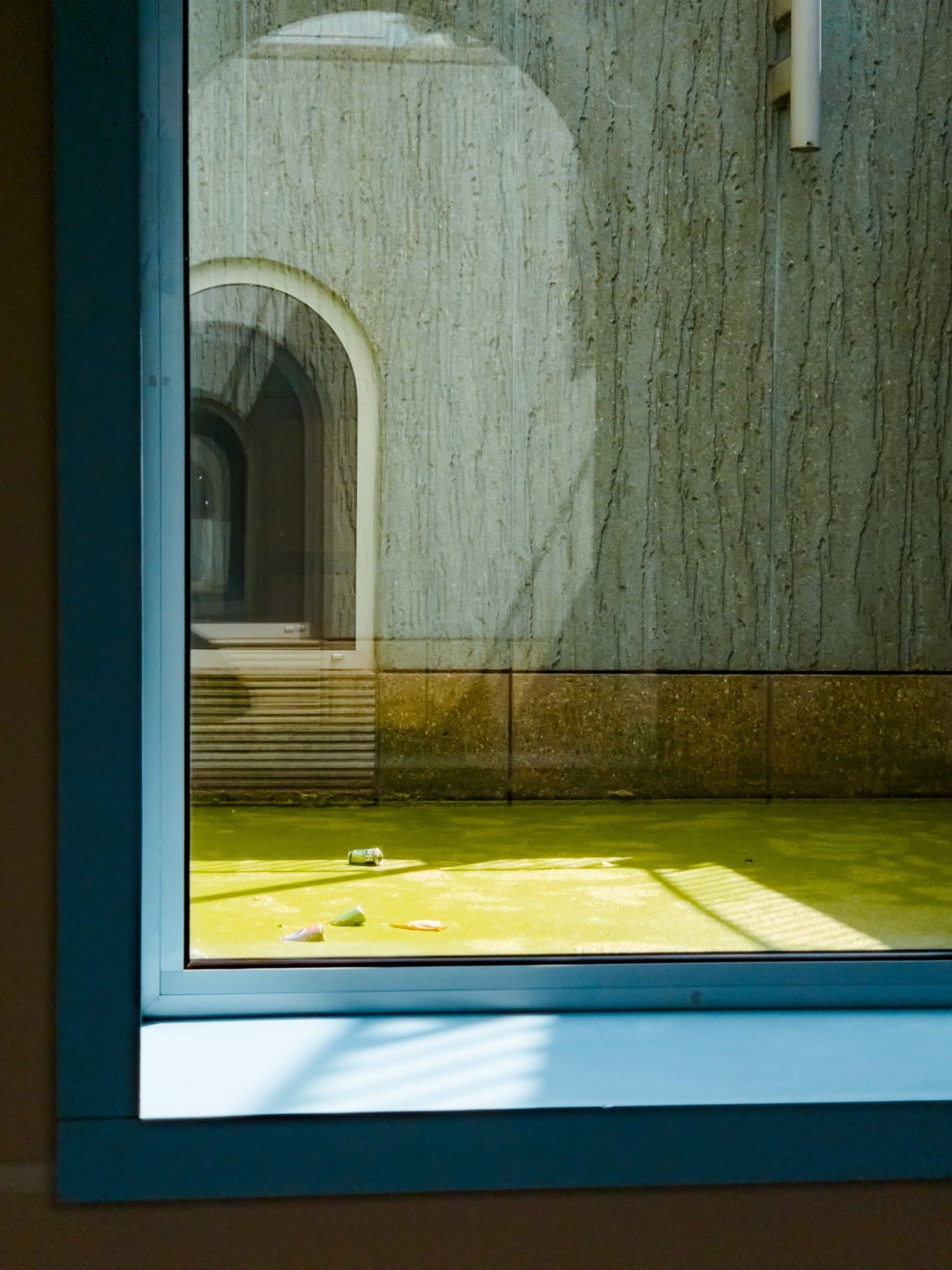 Doorkijkje door een raam in het Groninger Museum waar een stukje gracht bedekt met eendenkroos te zien is. Erop drijven lege blikjes bier. Aan de overkant is een betonnen gevel met eenzelfde boograam te zien van het museumgebouw.