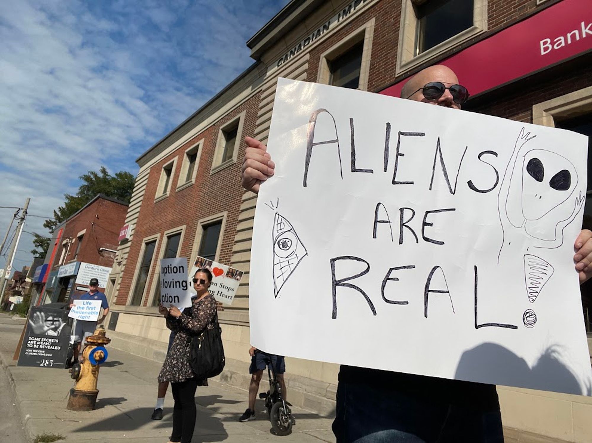 Me holding a hand-drawn poster with the text "Aliens are real" and a crude drawing of an alien with big eyes, and some grumpy people with anti-abortion signs behind me on the sidewalk