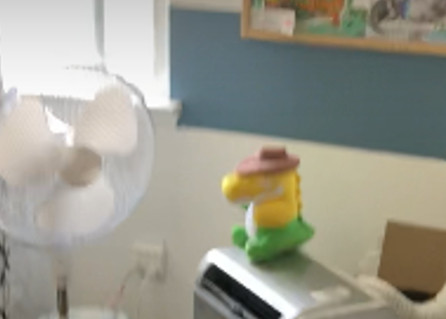 a low resolution photo of a smiling neon green and yellow alligator plushie wearing a cowboy hat sitting ontop of an air conditioner in front of a fan