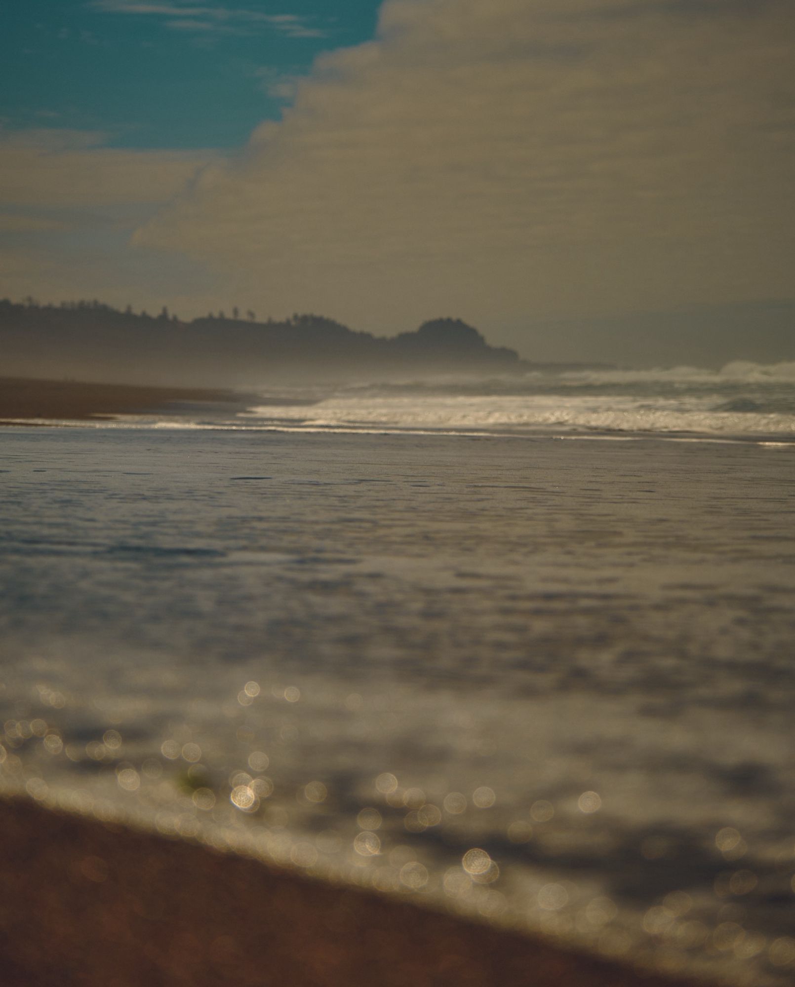 A color photo taken from a low angle along the Pacific Ocean, the incoming water blurred in the foreground, and the peninsula and waves are clear in the distance.