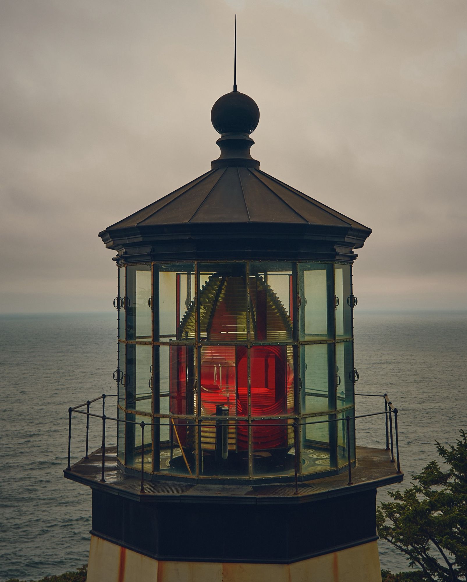 A color photo of the top of Cape Meares Lighthouse in Oregon. Its black iron facade and white tower show visible signs of weathering. The giant light housed inside is red with clear panels. The Pacific Ocean makes up the backdrop with a few pieces of foliage peeking in.