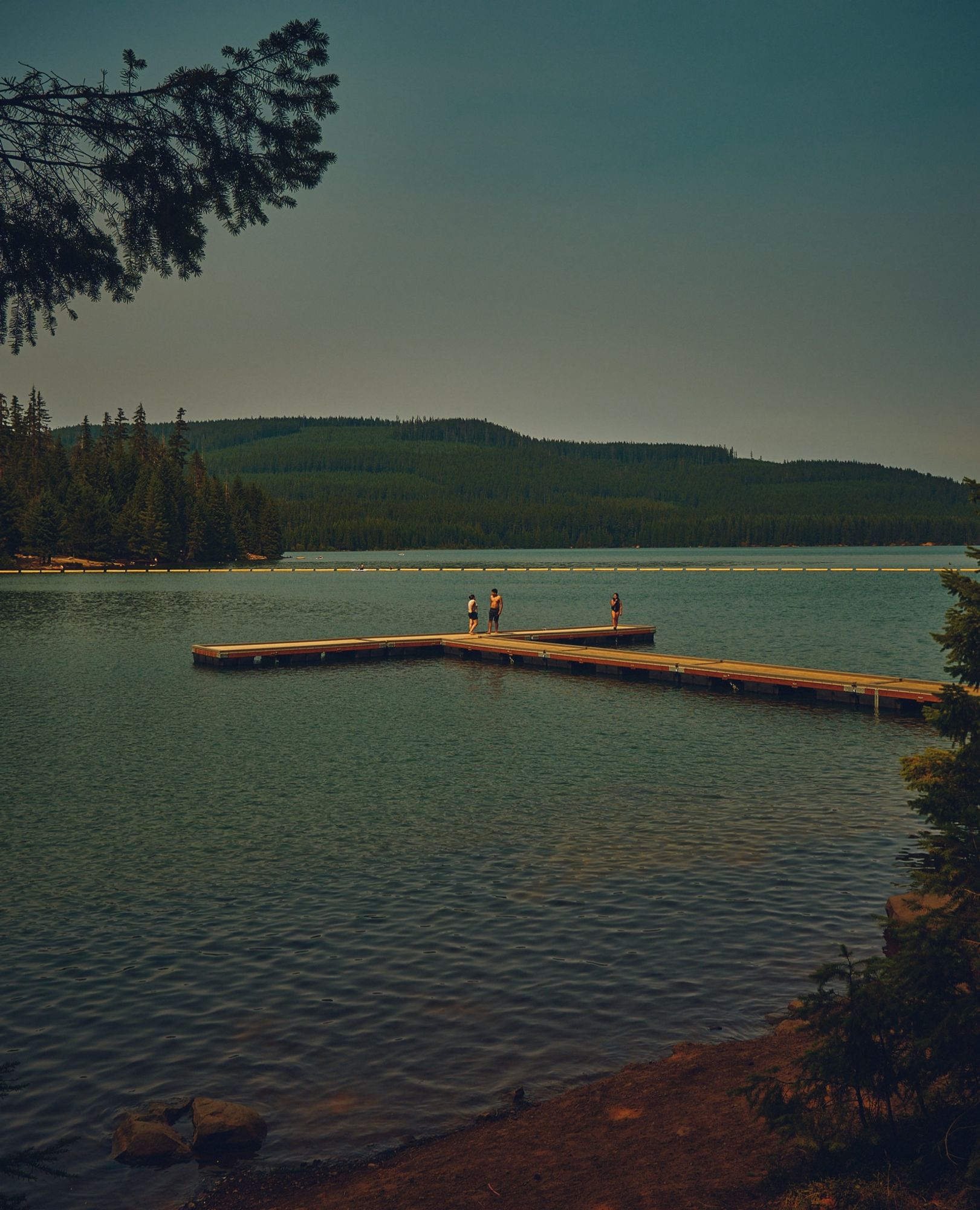 A color photo of kids standing on the end of an empty dock on a lake on a late summer evening.