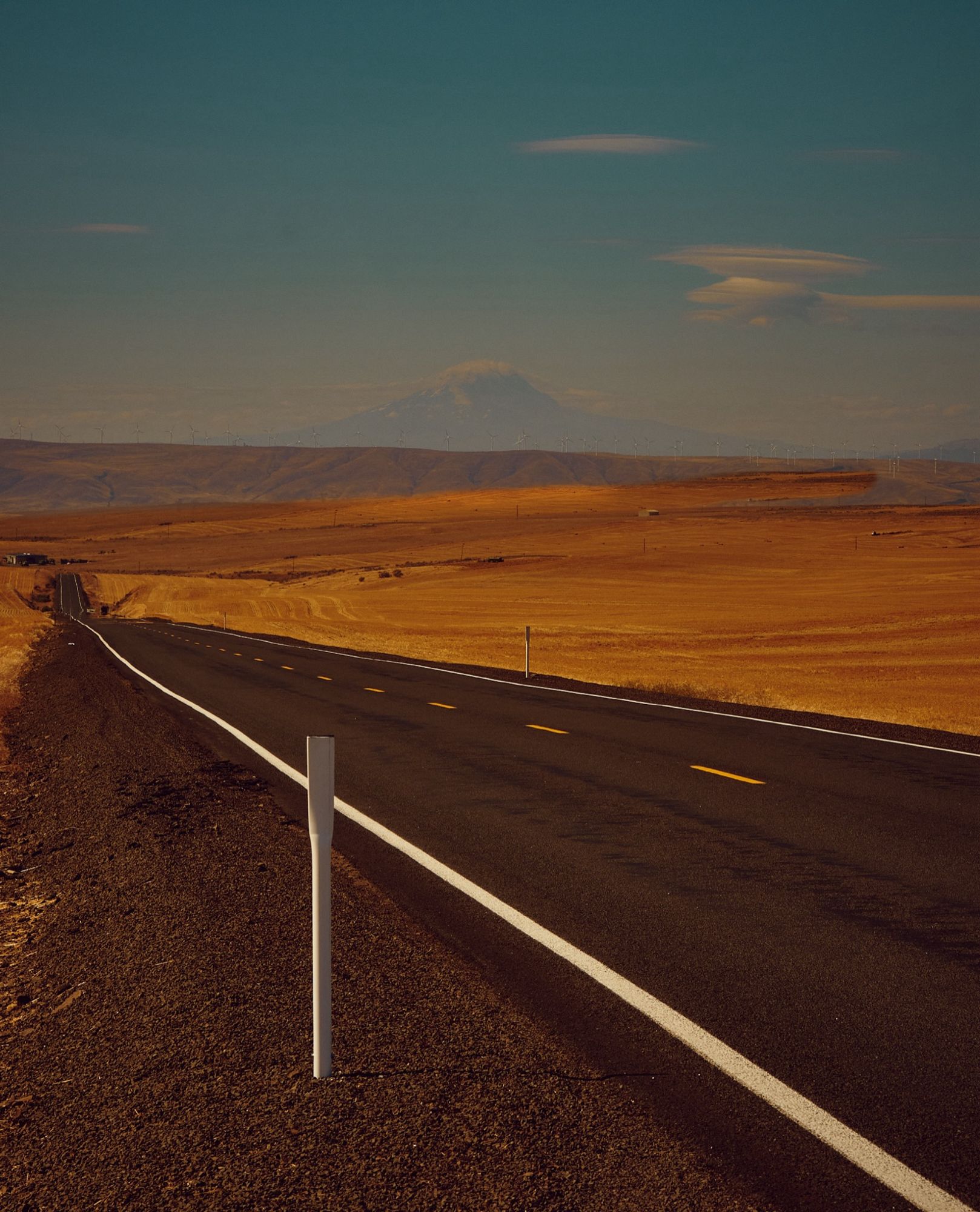 A color photo of an empty roadway leading to the hills among fields of dry grass.