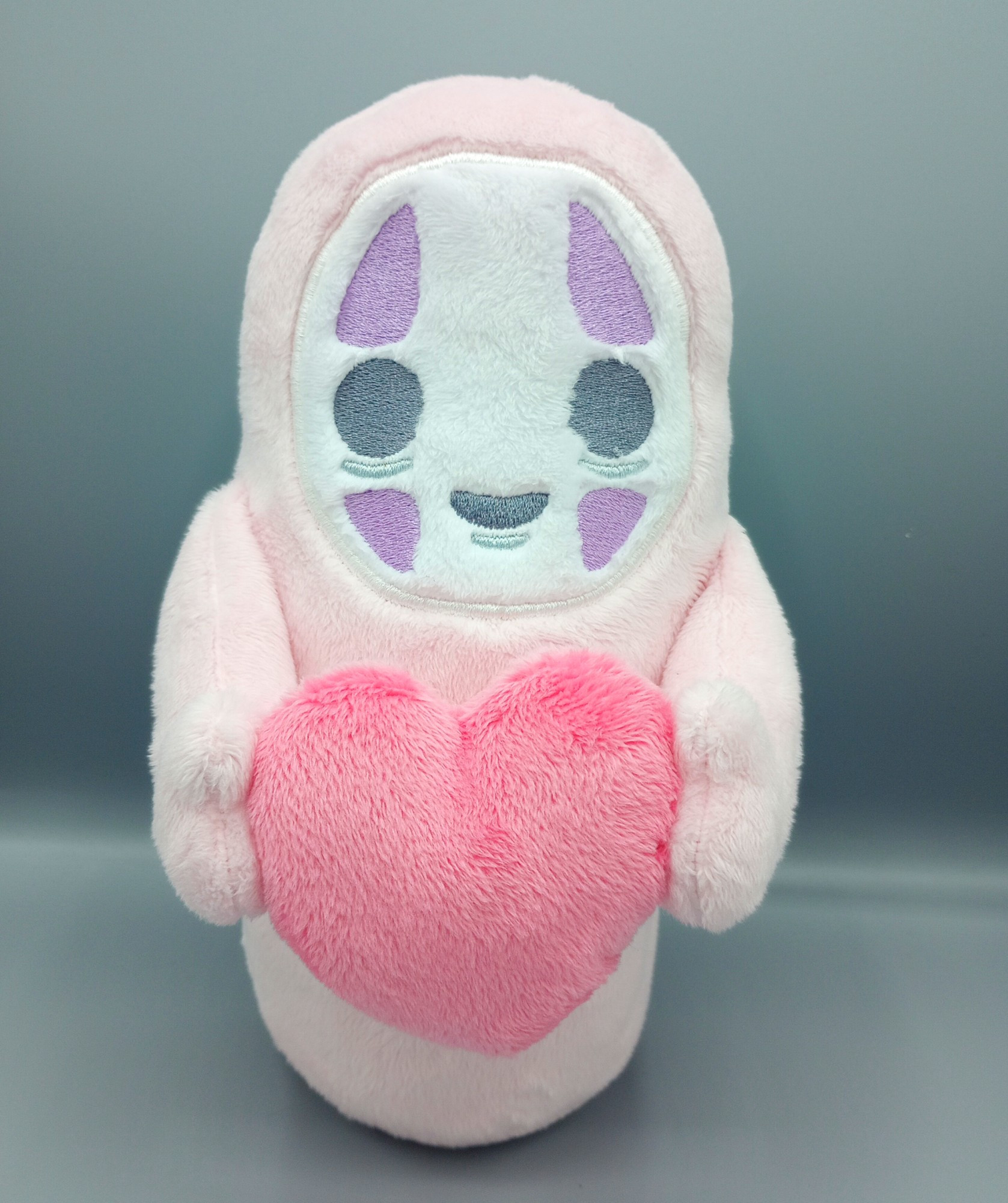 A handmade pink No Face plushie from the Studio Ghibli movie Spirited Away.  He is holding a pink heart.