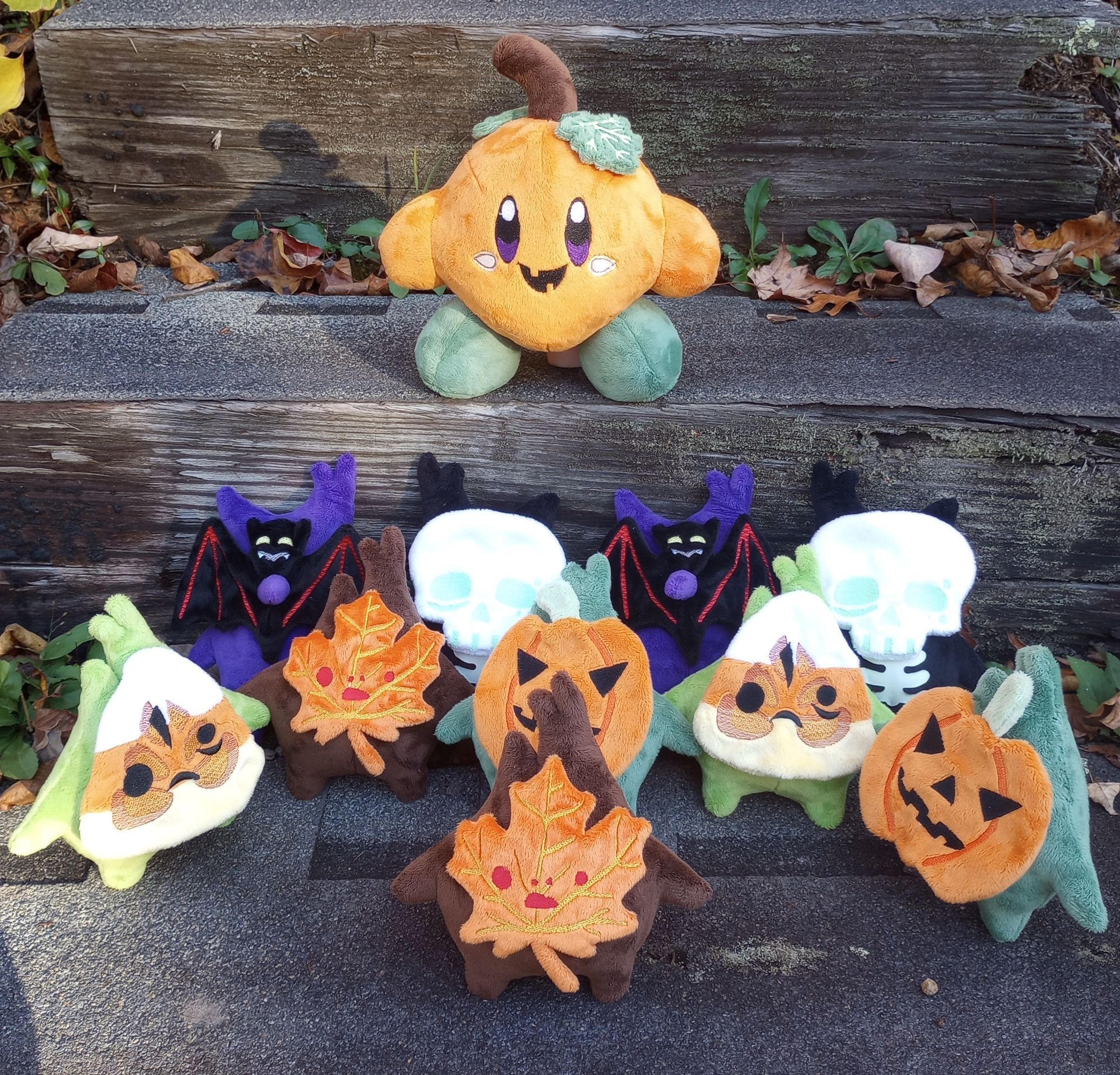 A collection of handmade Halloween plushies stands on some outdoor steps. The words "Halloween Plush Drop!" are above them.  On the top step is a Pumpkin Kirby plush, and on the step below are Candy Corn, Vampire Bat, Skeleton, Maple Leaf, and Pumpkin Korok plushies.