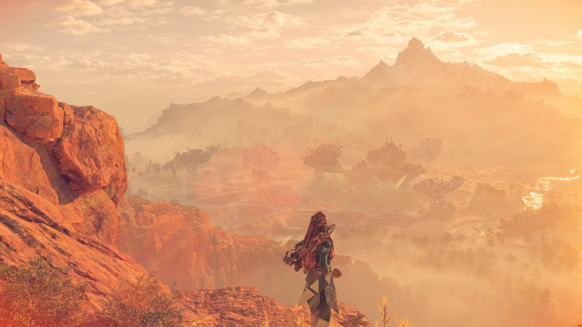 A screenshot from the animated action RPG “Horizon Forbidden West” - The player character, Aloy, stands on a mountain gazing over the horizon at sunset with mountains and trees in the distance. 