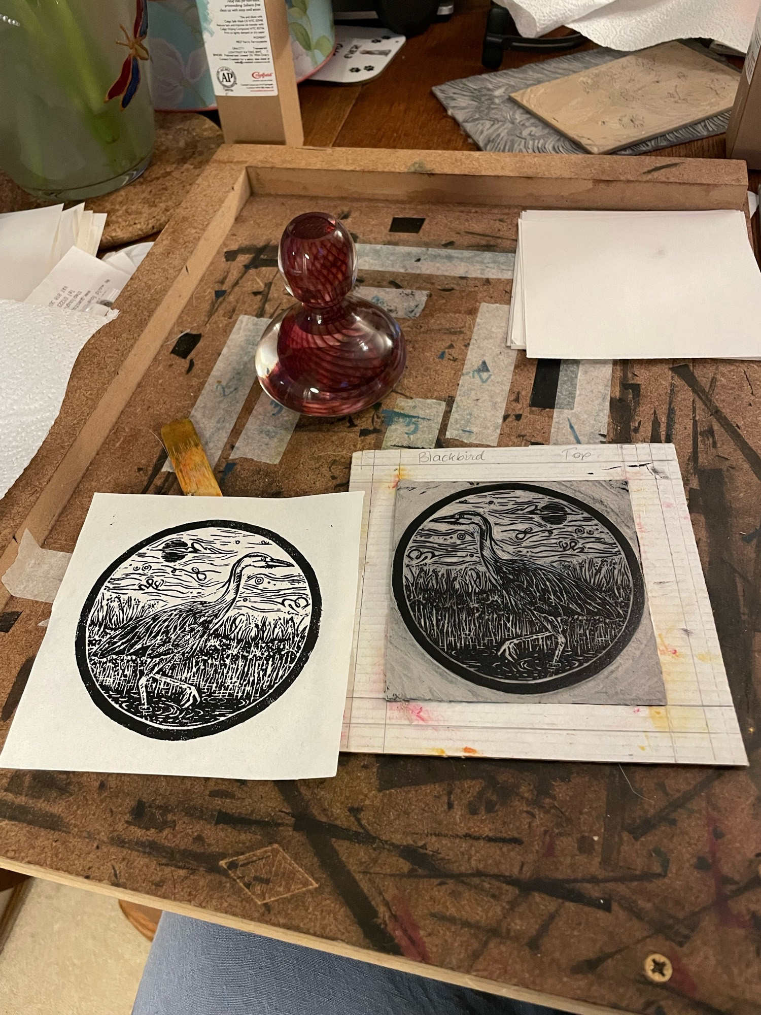 An inked up lino block of a heron standing in the reeds sits alongside a finished print. Also on the bench is a red swirled glass printing baren, it looks like a paperweight with a handle.