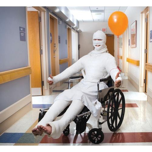 A person is sitting in a wheelchair with a whole body cast on. They're holding two thumbs up, and an orange balloon is tied to their left arm.